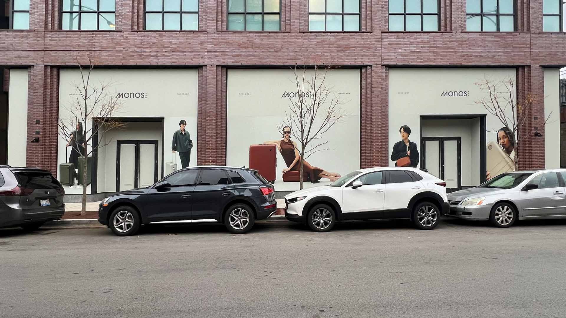 A street-level view of a brick building with store-front windows displaying advertisements, parked cars in the foreground.