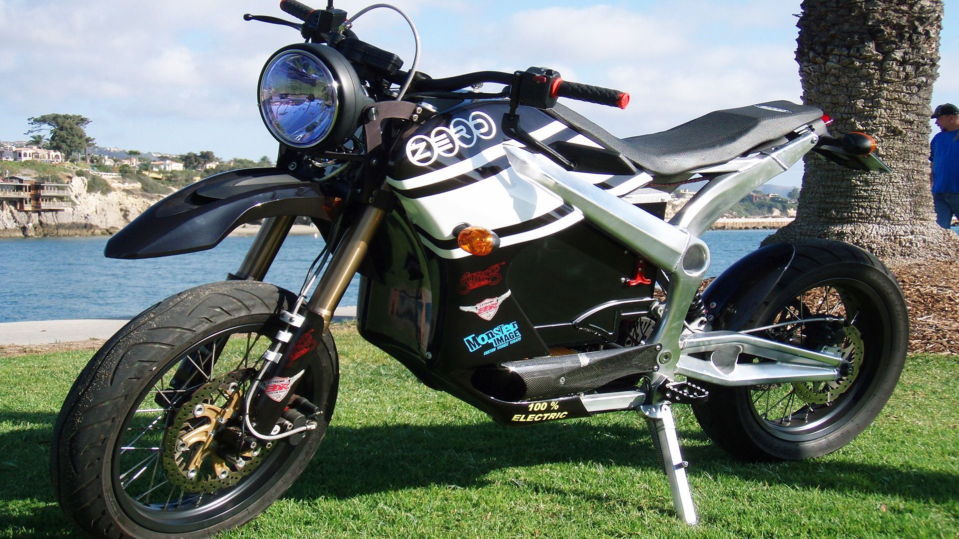 A black and white electric motorcycle parked on grass near a body of water, featuring gold-colored wheel hubs.