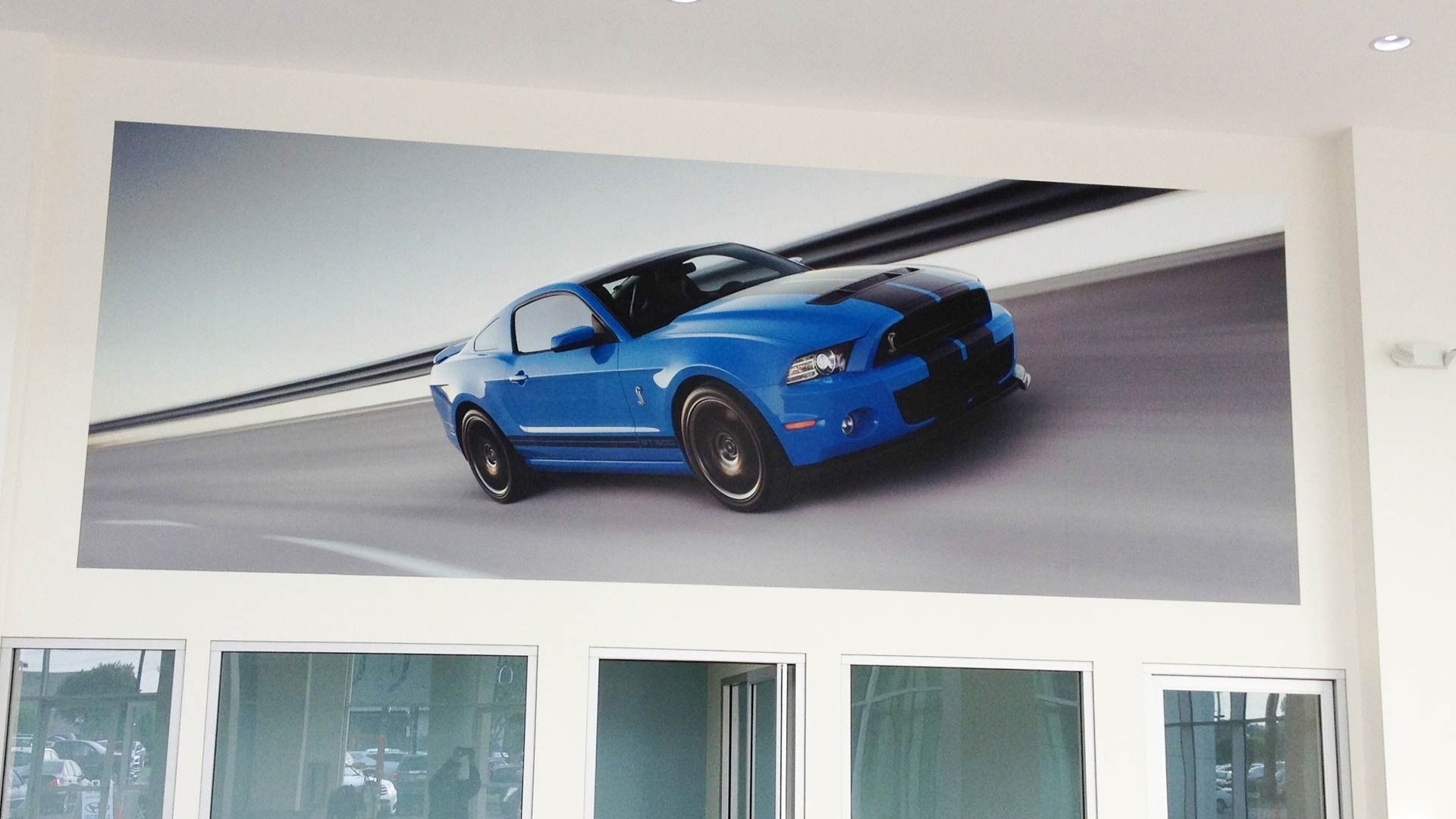 A large wall mural of a blue Ford Mustang driving on a highway, mounted above a series of glass windows.