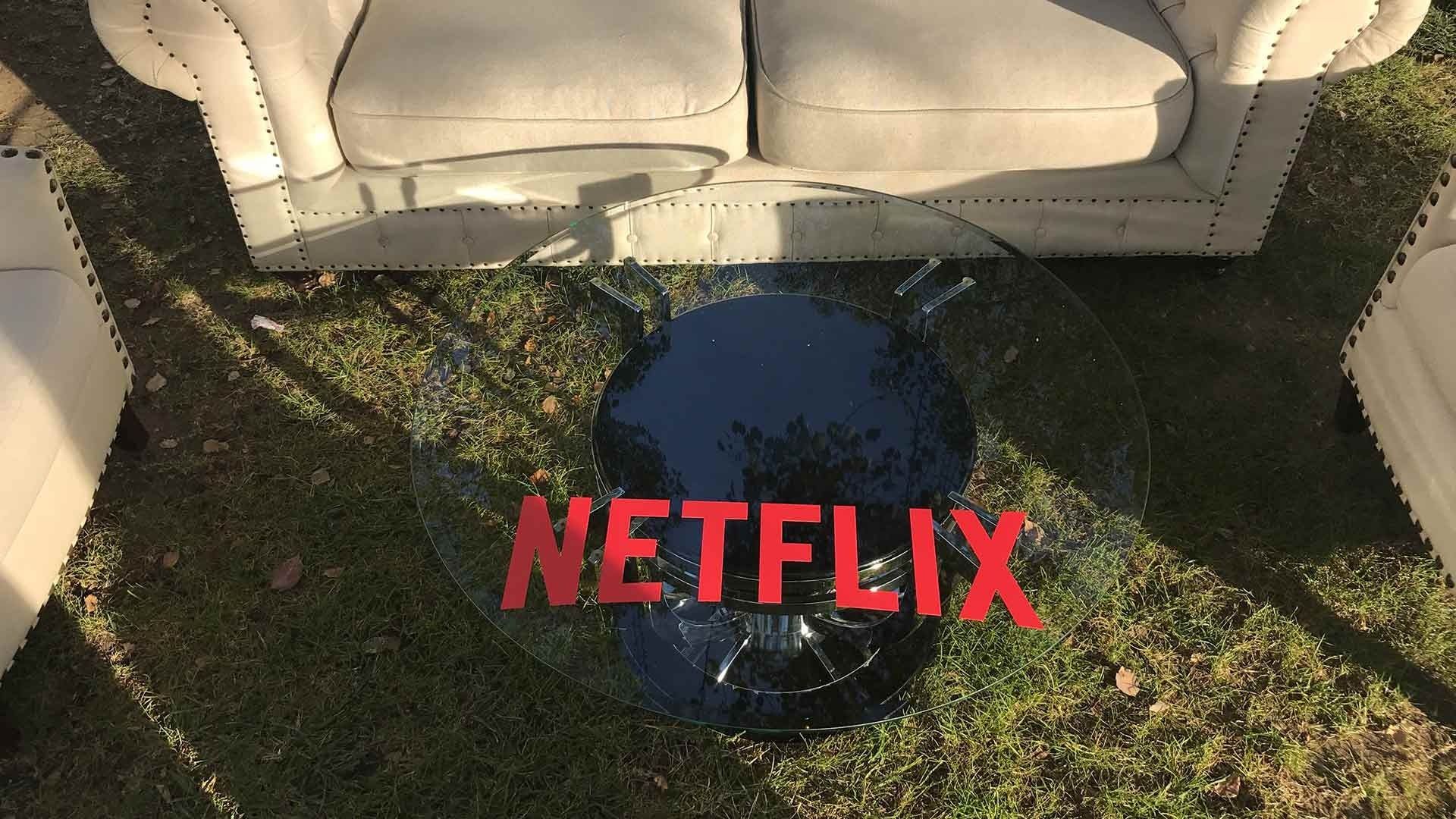 A round glass coffee table with the red Netflix logo sits on a grassy lawn in front of beige cushioned sofas.
