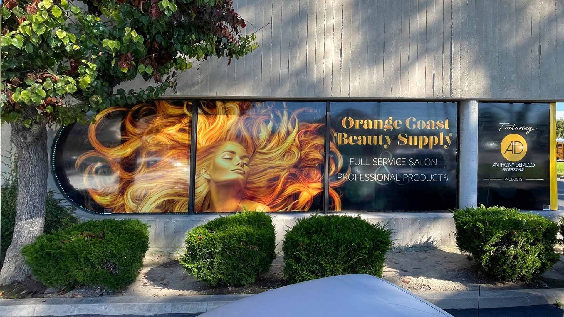 Orange Coast Beauty Supply storefront featuring a window graphic of flowing hair, with shrubs in the foreground.