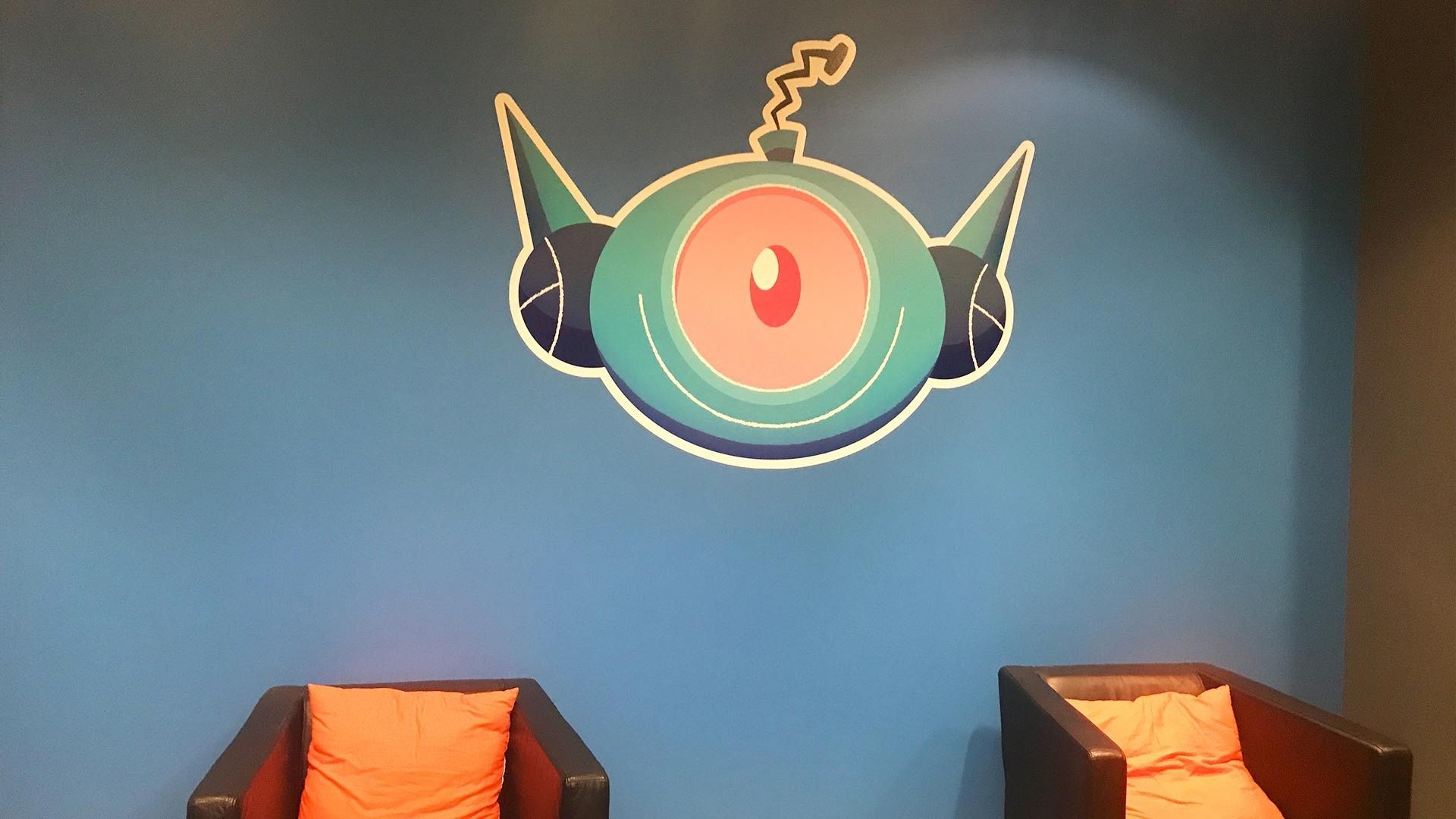 A blue wall features a graphic of a green, one-eyed, robotic-looking head, positioned above two chairs with orange cushions.