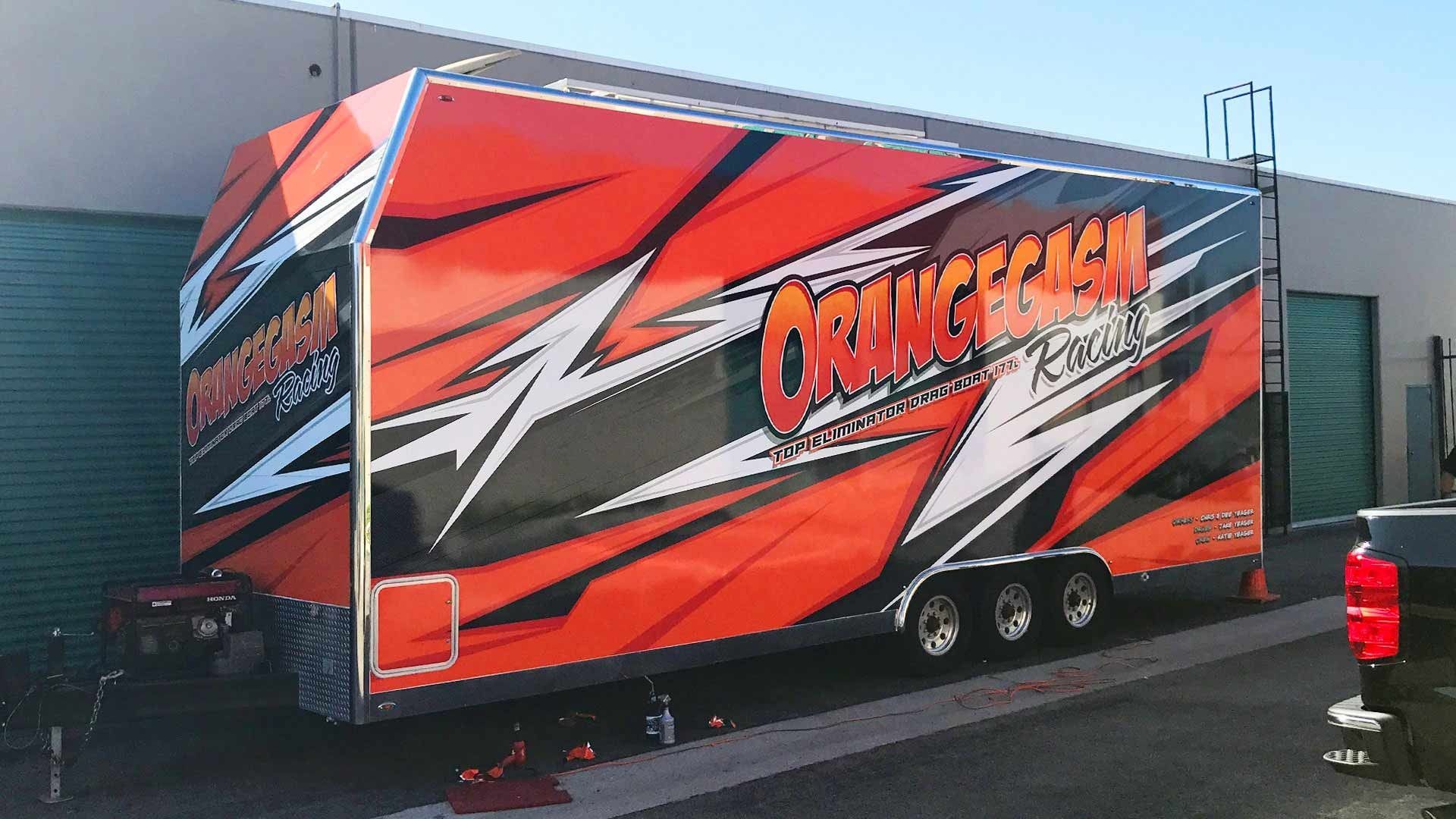 A large, triple-axle trailer with a bright orange and black graphic wrap parked next to a building on a sunny day.