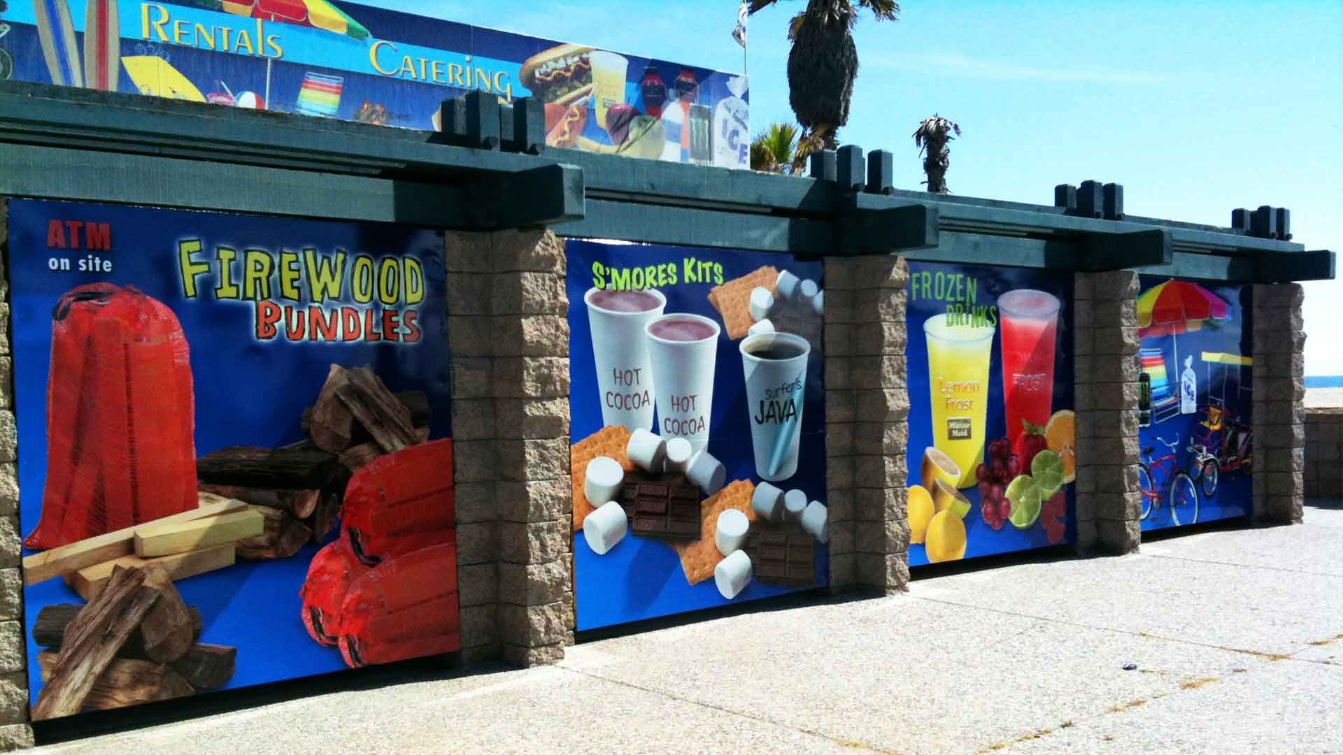 A row of beachside concession stands with blue signs advertising firewood, s'mores kits, and colorful frozen drinks.
