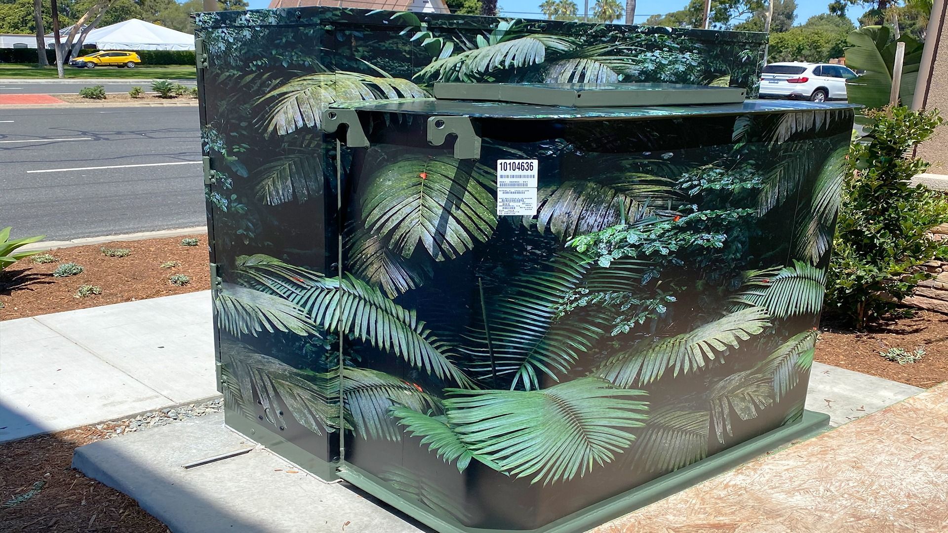 A metal utility box covered in a green and black fern leaf pattern, located on a sidewalk near a parking lot.