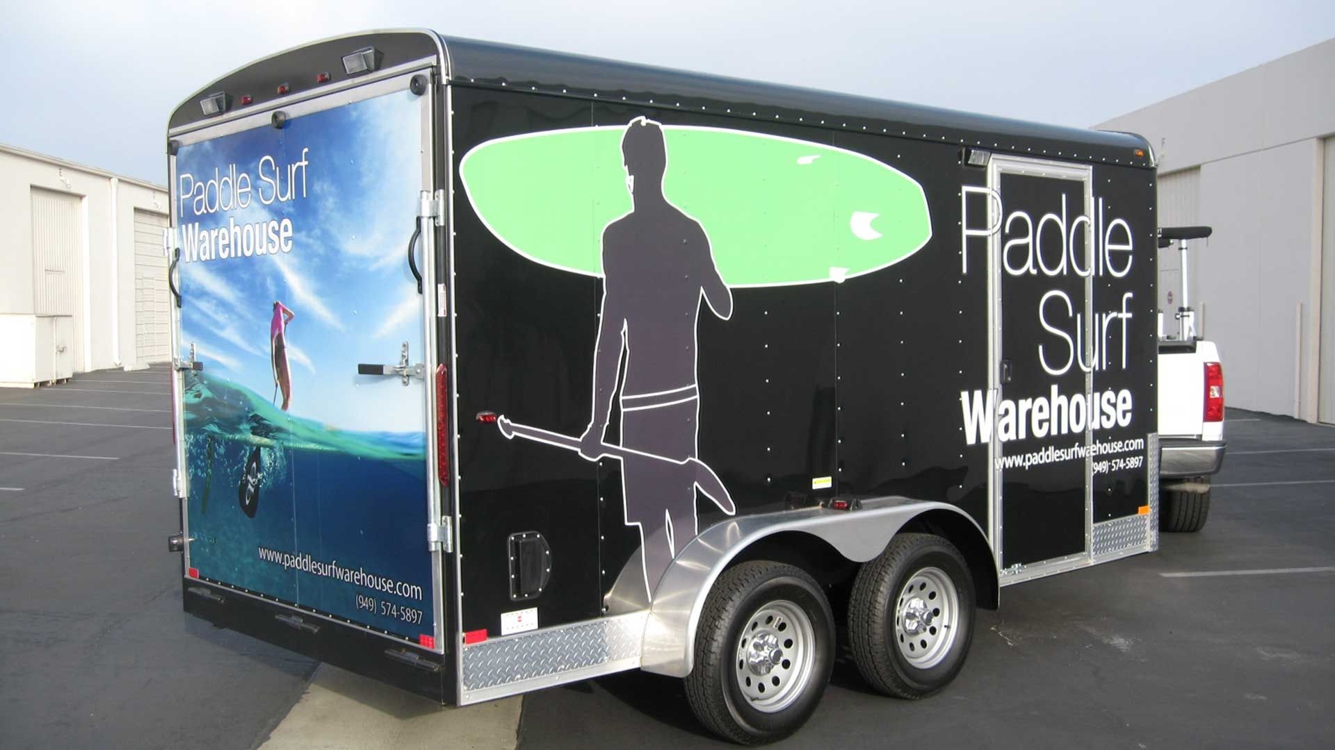 A black trailer parked in a lot, featuring Paddle Surf Warehouse branding and a silhouette of a person with a surfboard.
