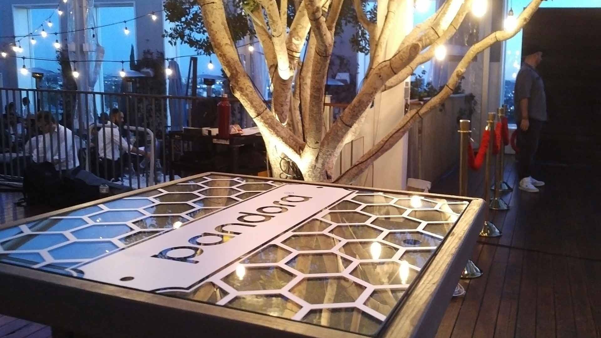 A Pandora-branded table with a honeycomb design sits outdoors under string lights next to a tree, with people nearby.