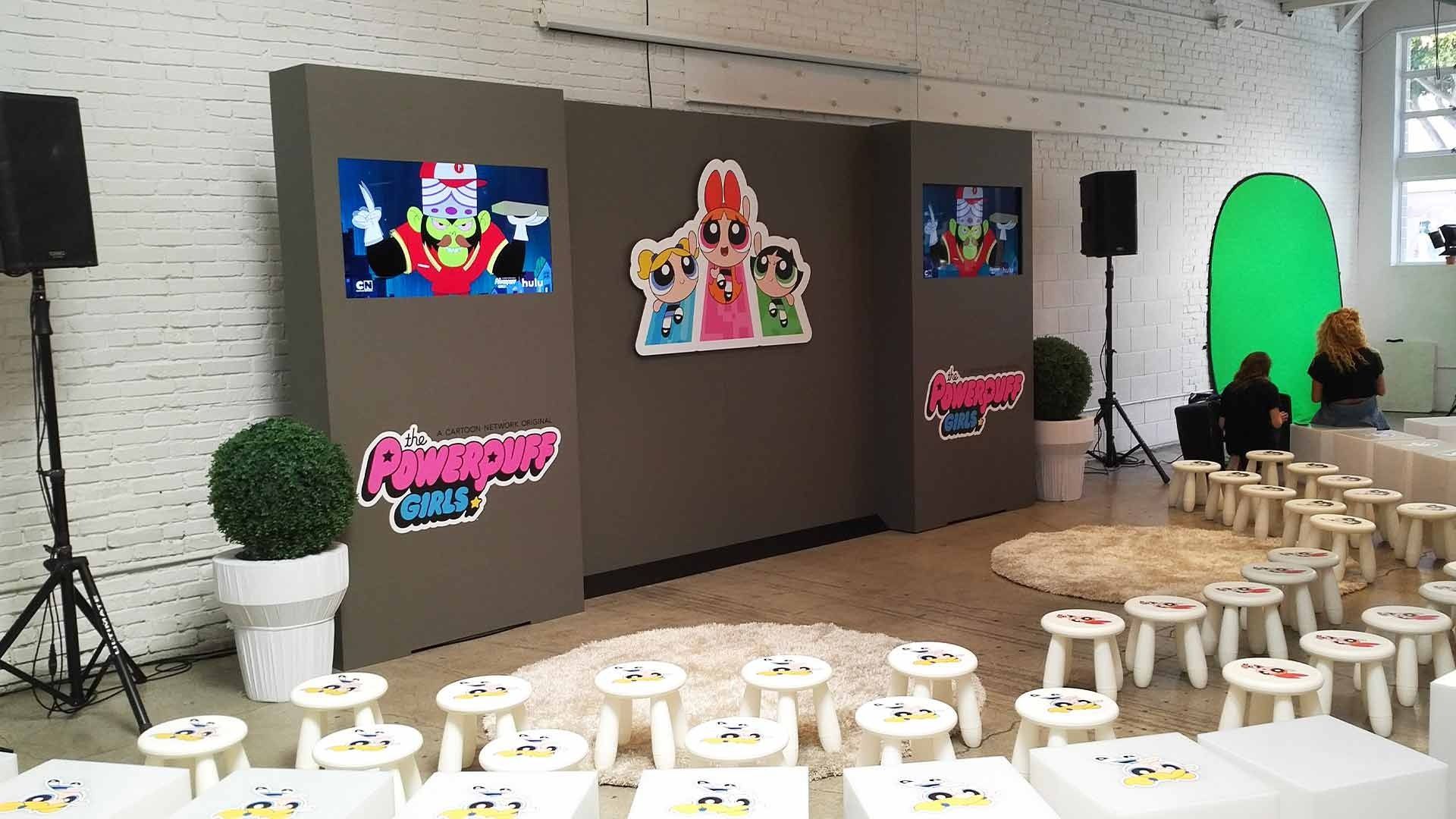 A Powerpuff Girls-themed event space with a center display, stools for attendees, and a green screen area.
