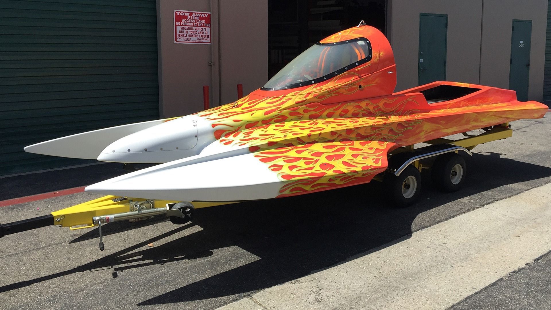 A yellow, orange, and white racing boat on a trailer parked outside a building.