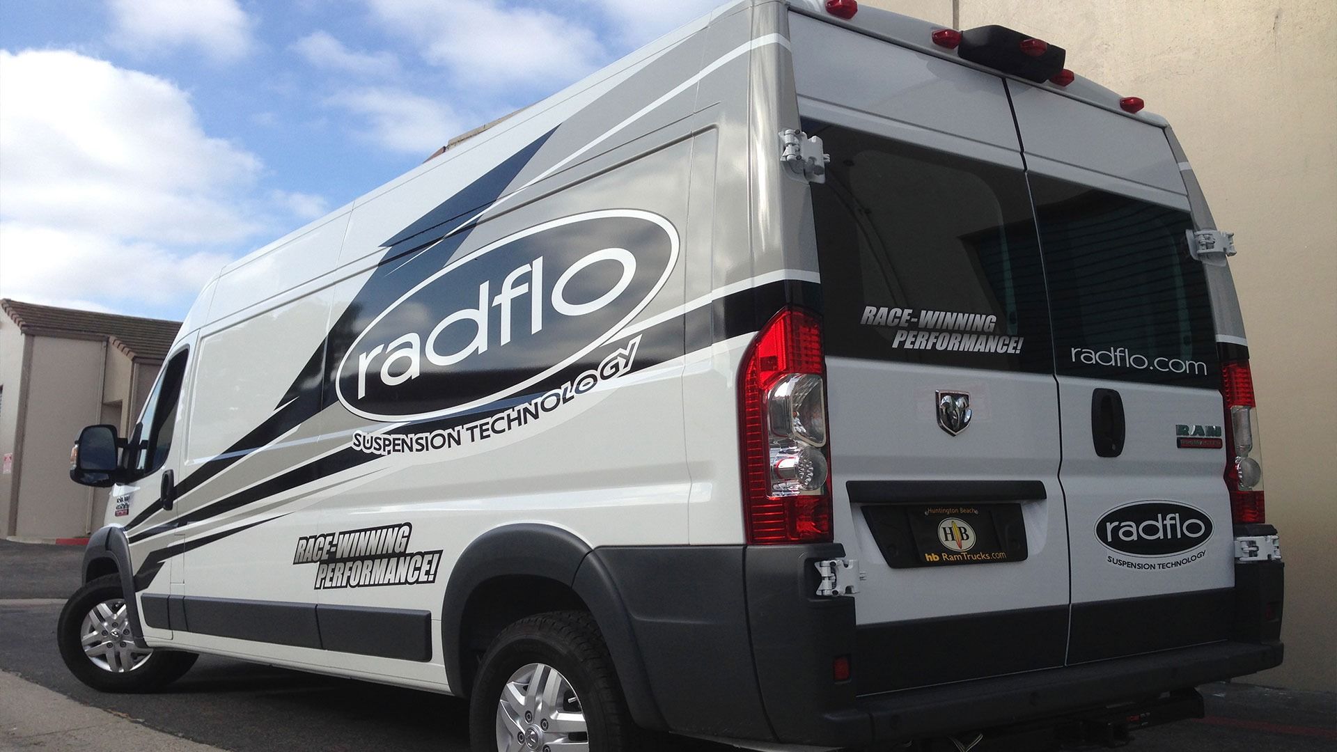 White Ram ProMaster van branded with Radflo suspension graphics, parked outside against a plain building wall.