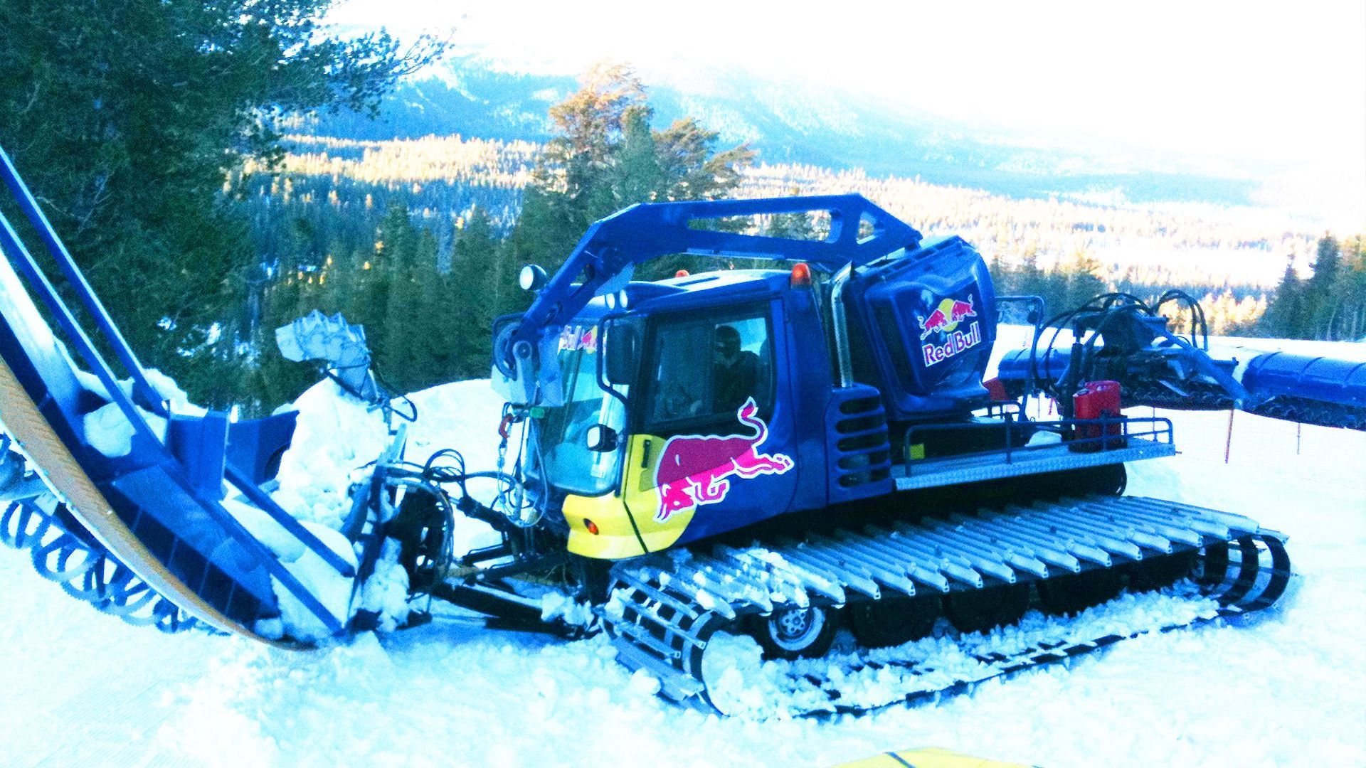 A blue Red Bull snow groomer parked on a snowy mountain slope with pine trees in the background.