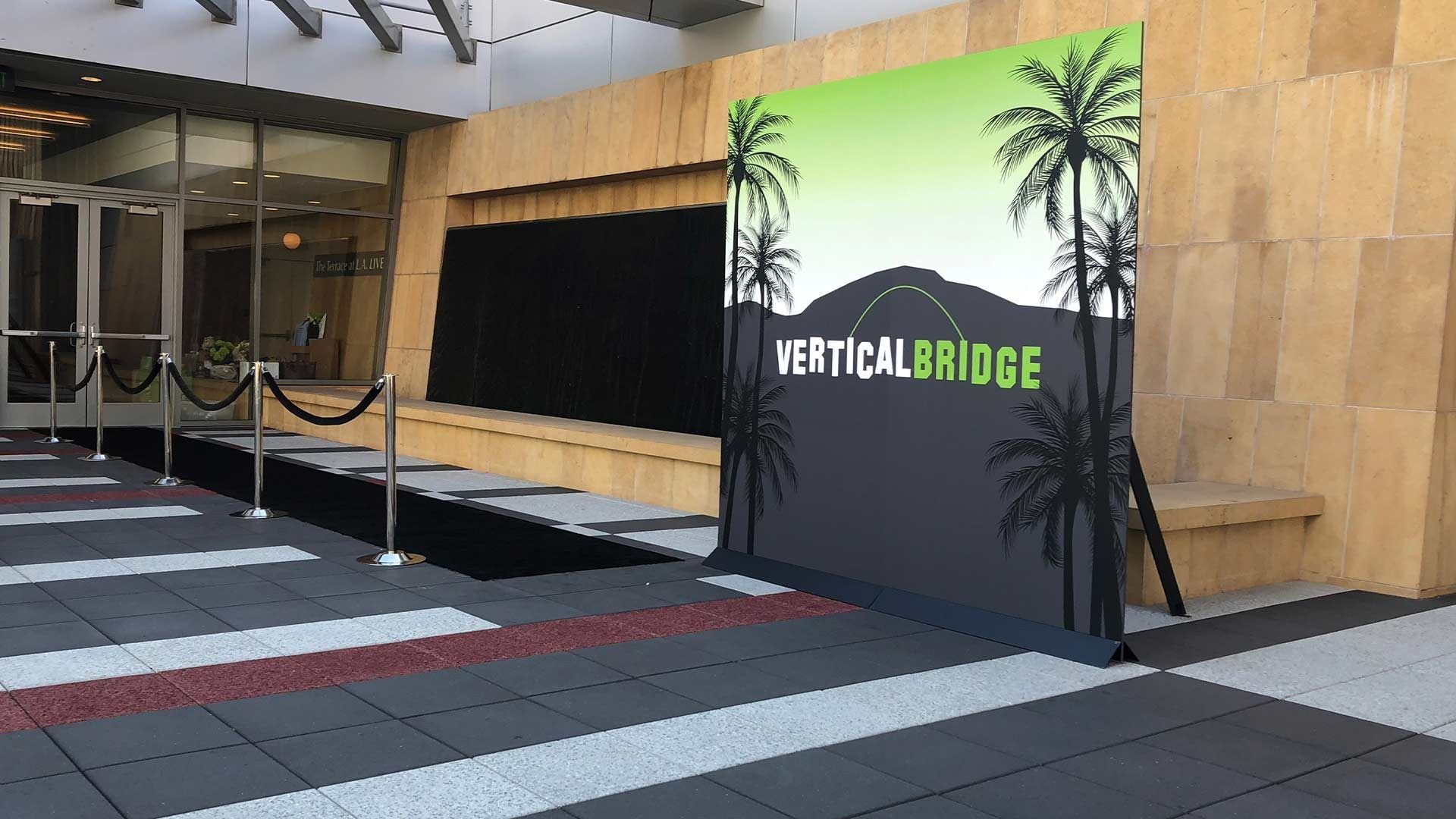 A branded step-and-repeat backdrop for Vertical Bridge stands outside a building with a red-carpet-style entrance.