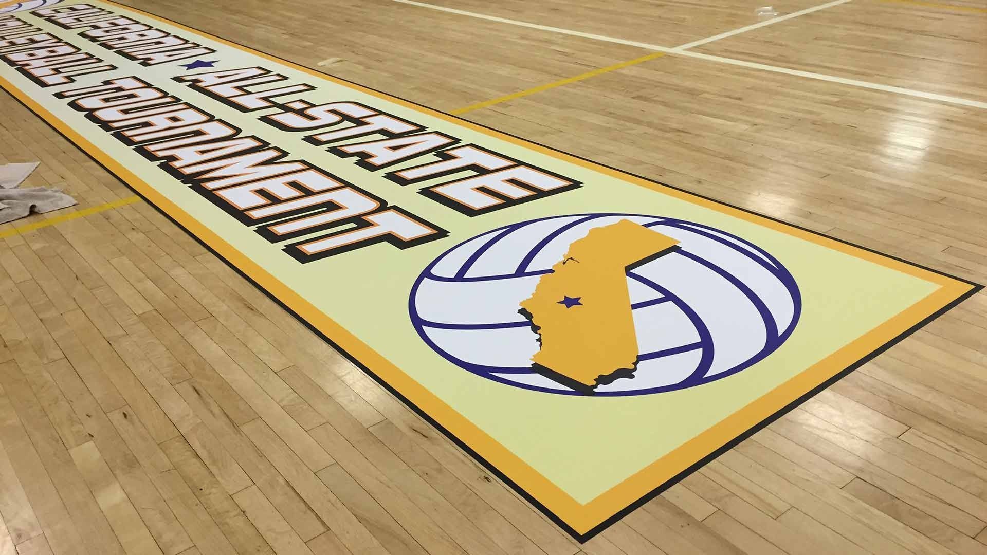 A volleyball tournament banner featuring a volleyball icon with an orange state outline, placed on a wooden gym floor.