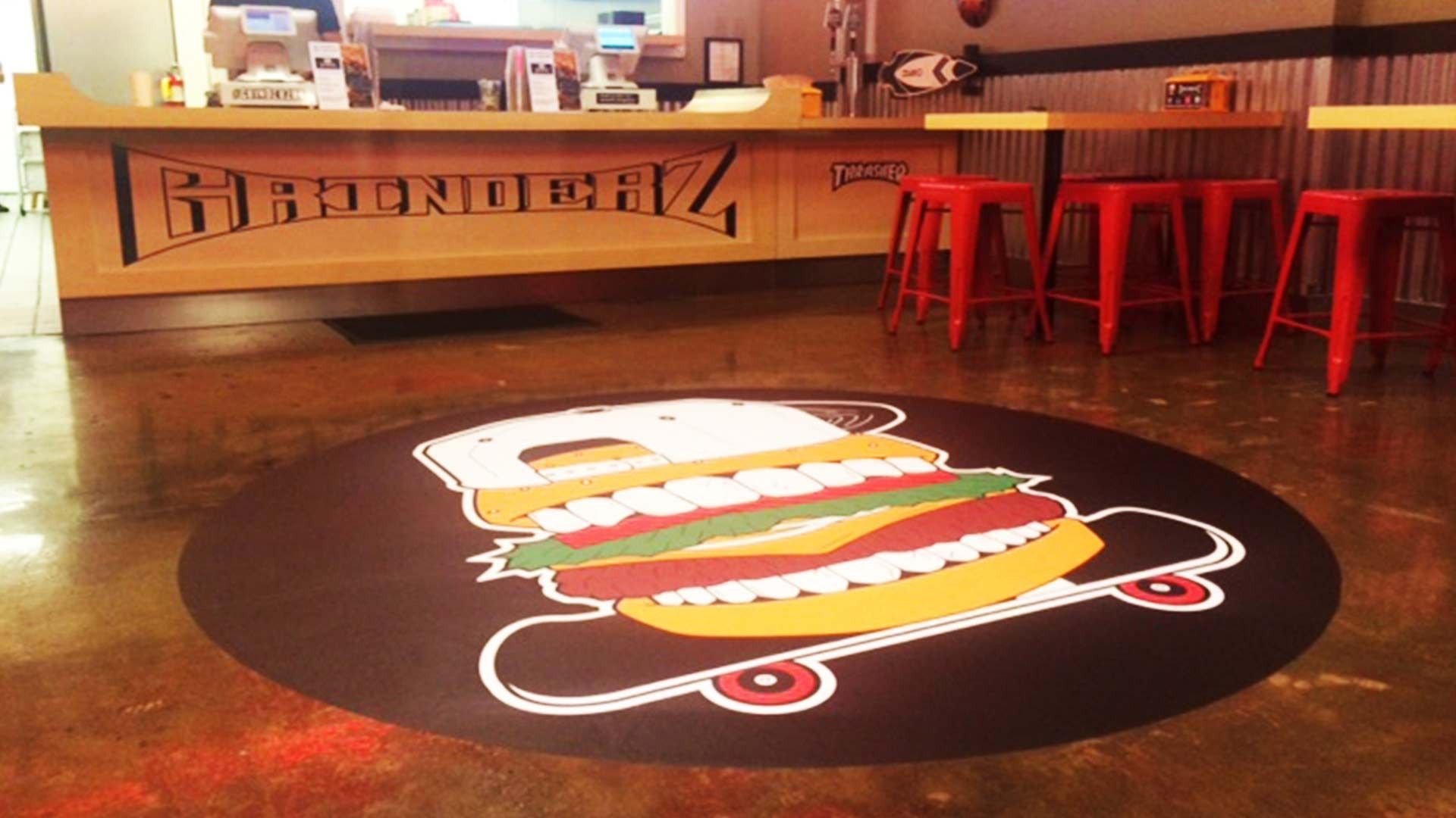 A round burger-on-a-skateboard decal on a concrete floor in front of a restaurant counter with red stools.