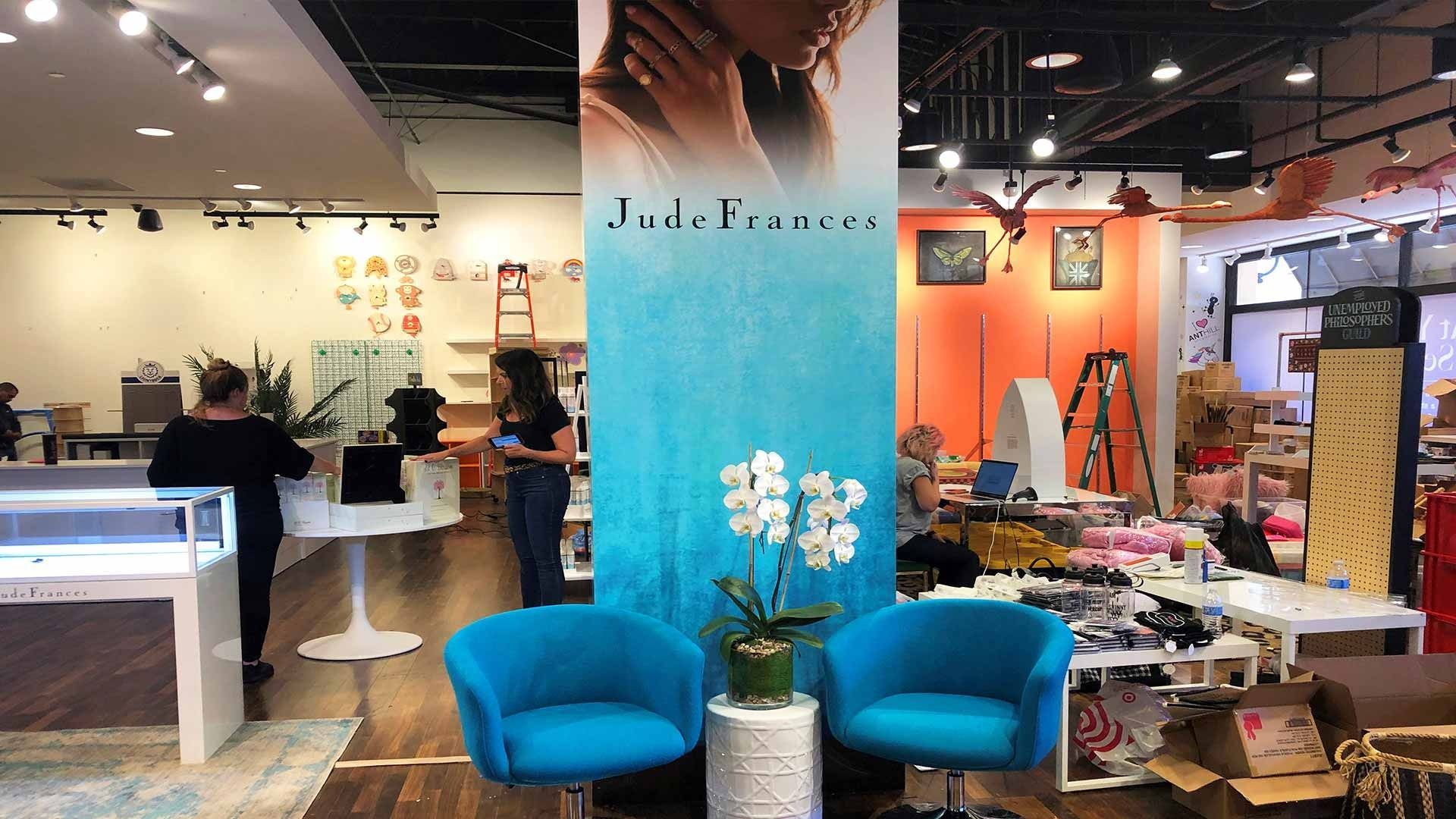 A retail jewelry store features a turquoise pillar with an advertisement, two teal chairs, and staff helping customers.