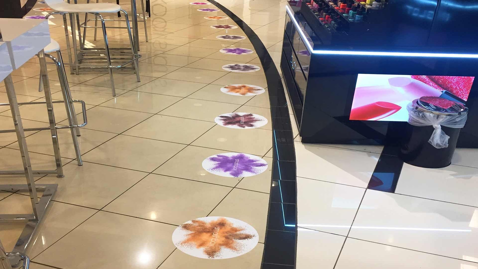 A row of colorful circular floor decals leads along a path next to a display case in a brightly lit, tiled retail store.