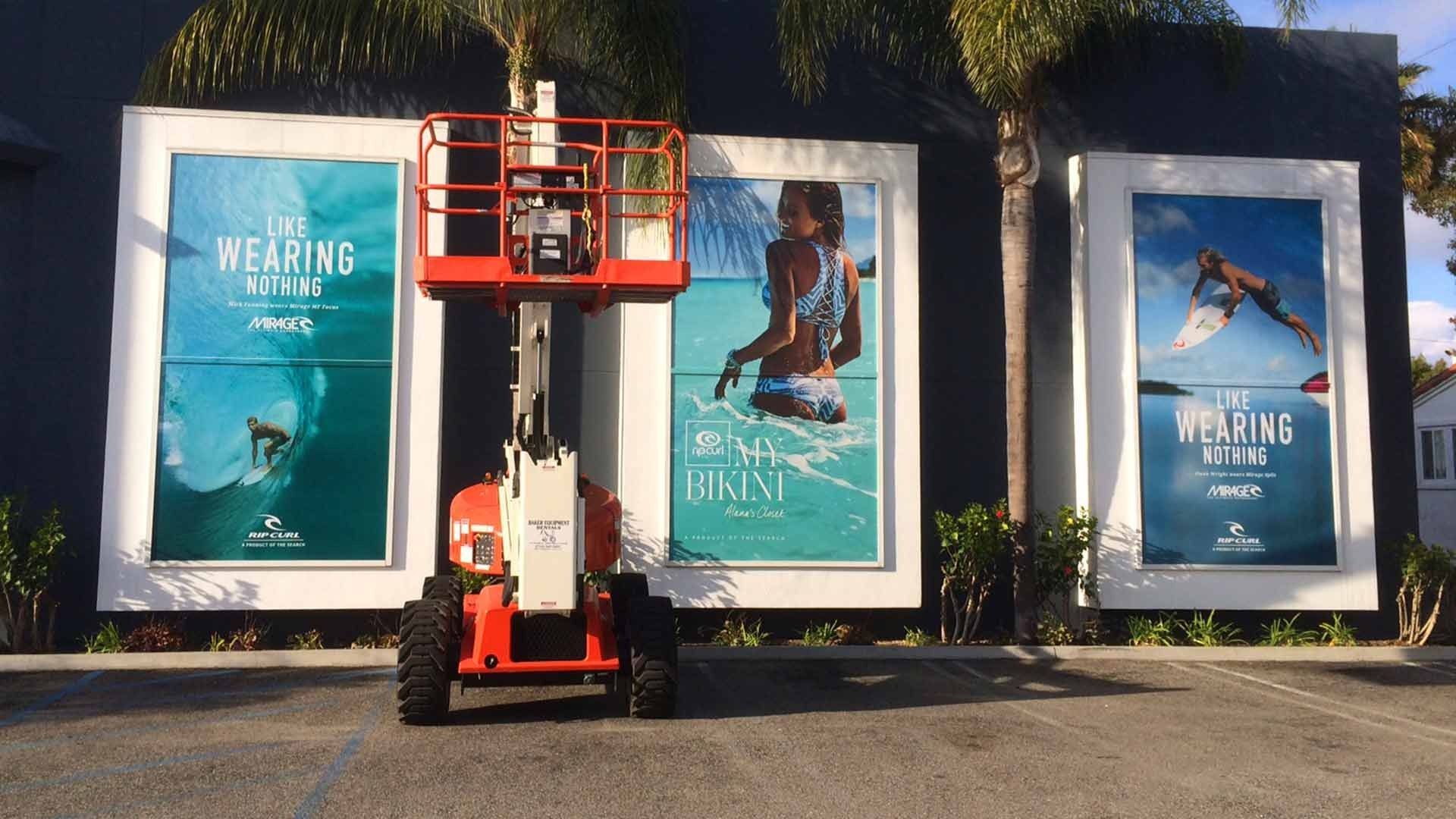 An orange scissor lift parked in front of a building with three large outdoor advertisements featuring beach scenes.