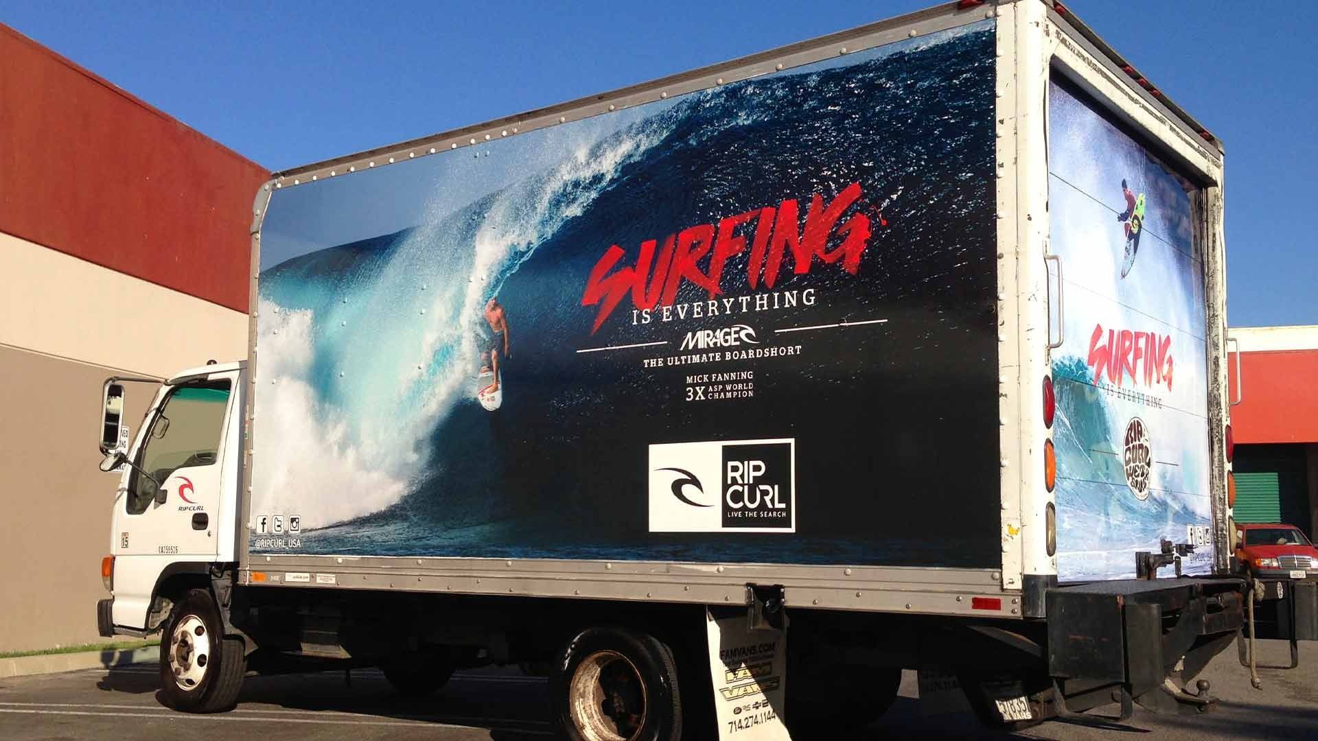A white box truck parked outdoors, featuring a large graphic of a surfer on a wave and Rip Curl branding on its side.