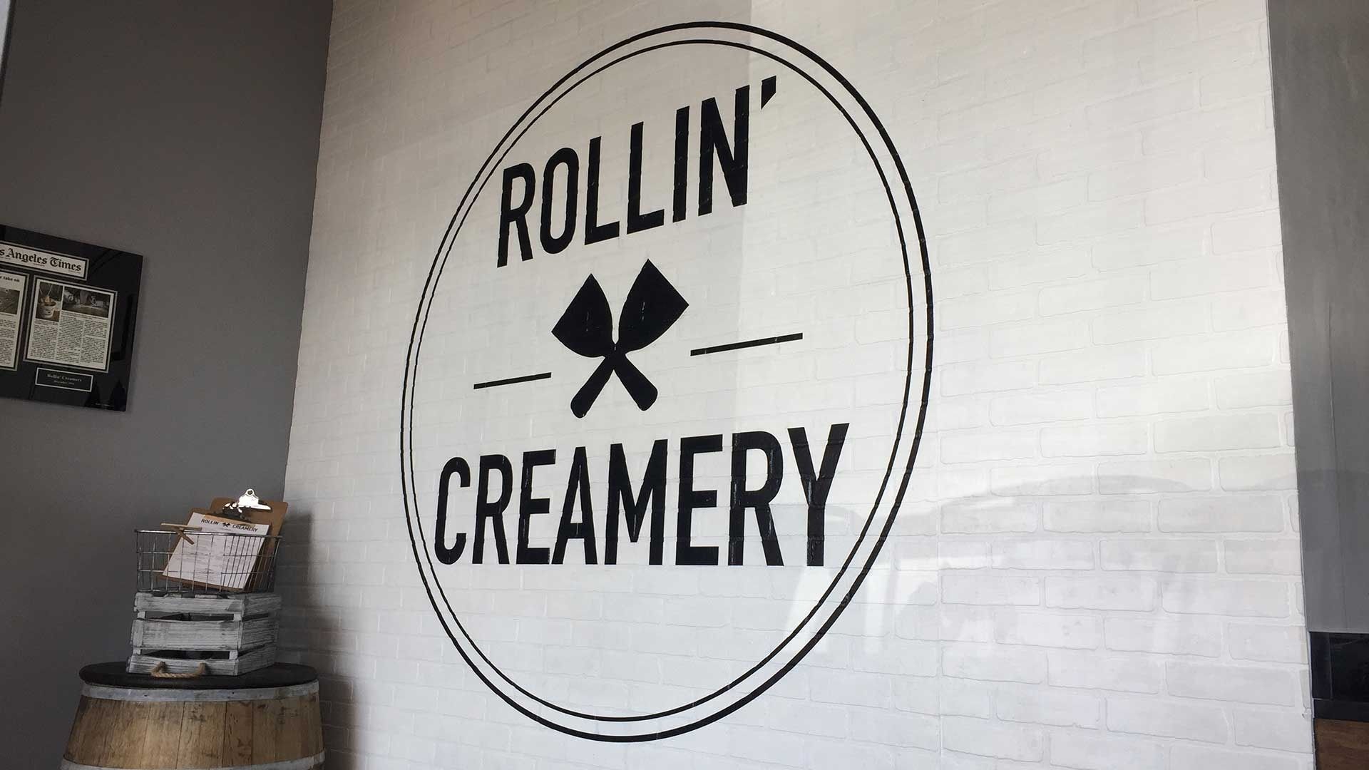 The Rollin' Creamery logo in black text with a crossed spatulas icon painted on a white brick wall.