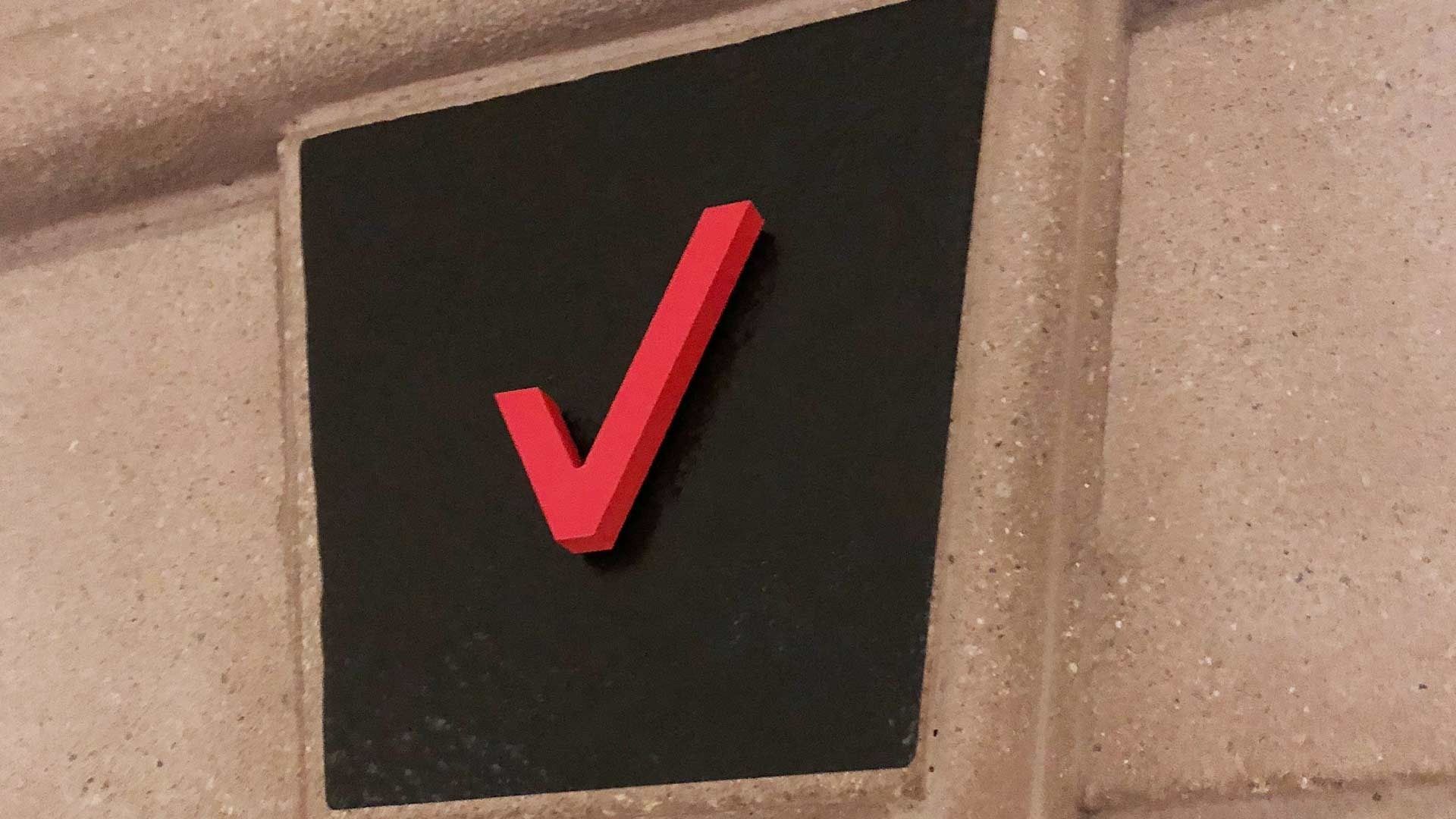 A bright red, three-dimensional checkmark mounted on a black square background, set against a light-colored stone wall.