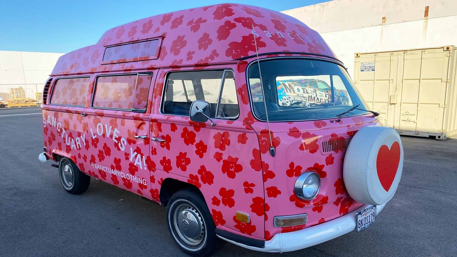 A pink Volkswagen van covered in red flower patterns, parked outdoors with a heart emblem on its spare tire cover.