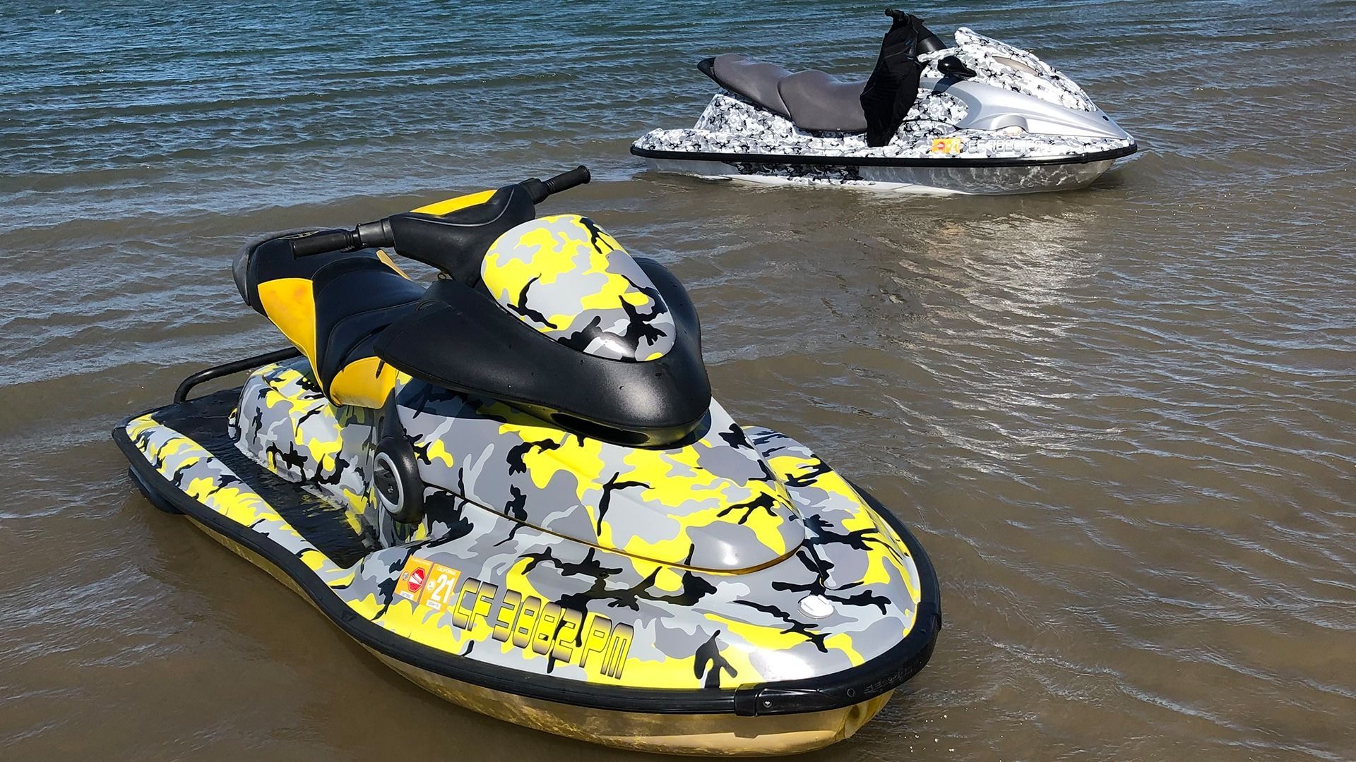 Two jet skis, one with yellow and gray camouflage and one with black and white camouflage, sitting in shallow water.