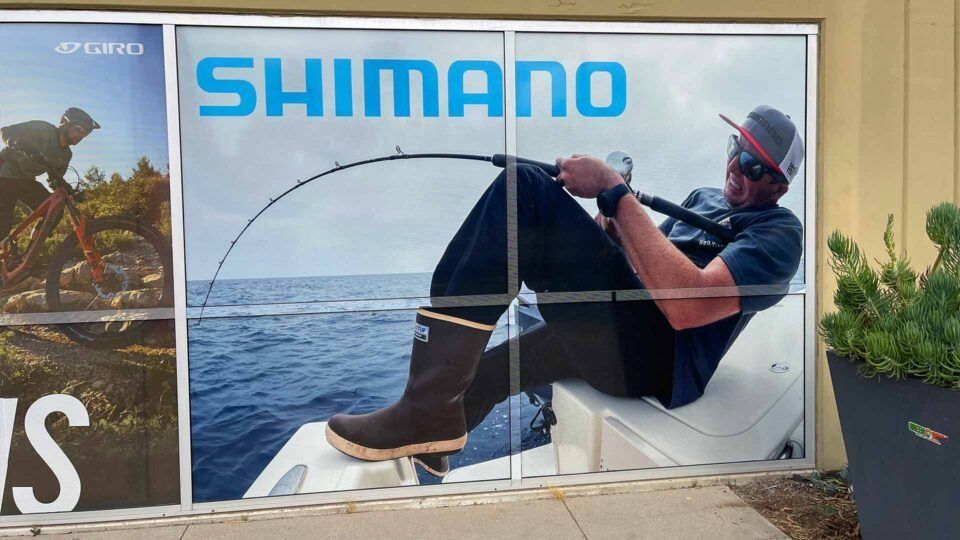 A vinyl store window display features a Shimano logo above a person fishing from a boat on the water.