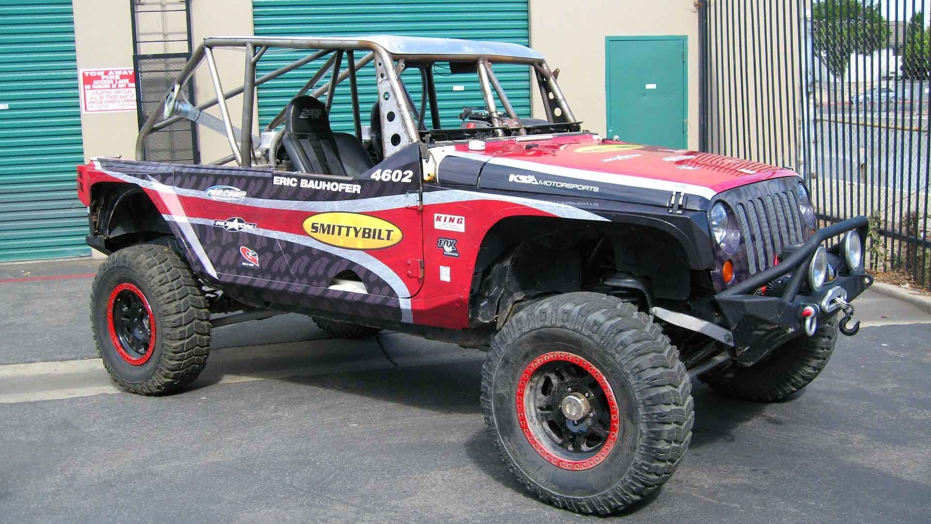 A custom red, black, and white off-road Jeep-style vehicle with a roll cage and large tires parked on a paved lot.