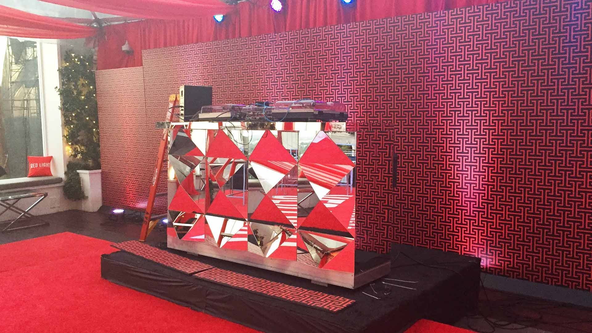 A DJ booth on a stage with a red patterned backdrop and a red carpet in a room with a decorated holiday tree.