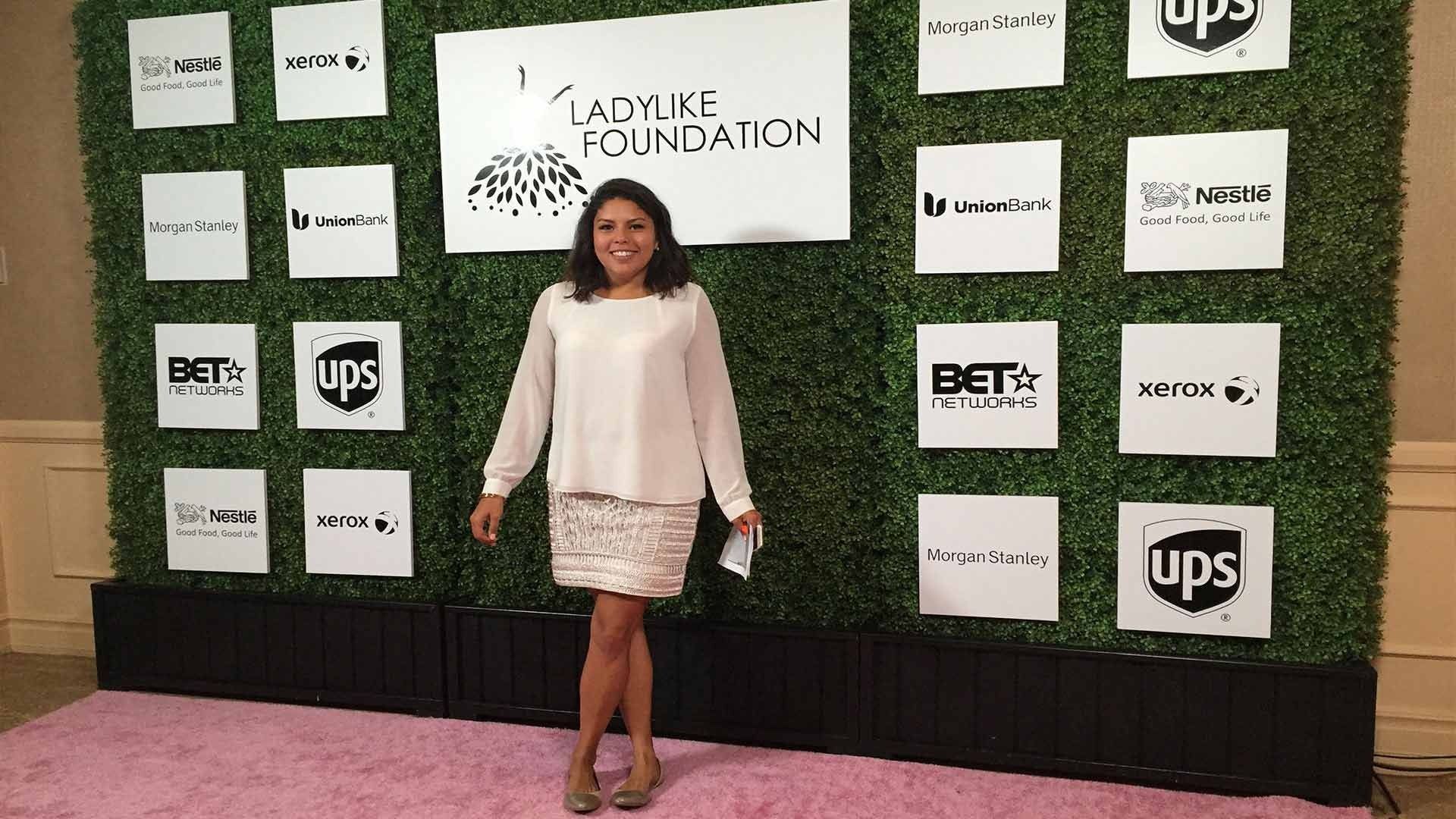 A person smiles while standing on a pink carpet in front of a green hedge backdrop with Ladylike Foundation logos.