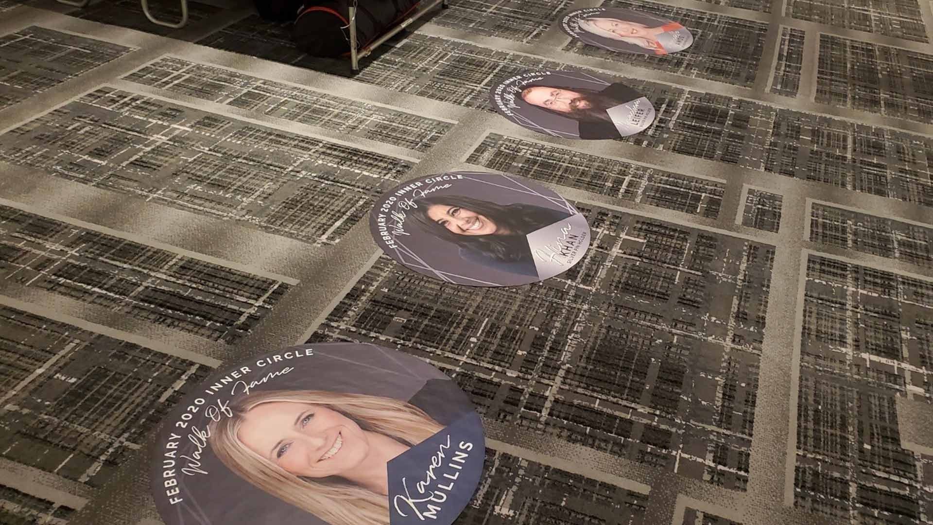 Circular floor decals featuring portraits of four individuals displayed in a line on a patterned carpeted hallway.