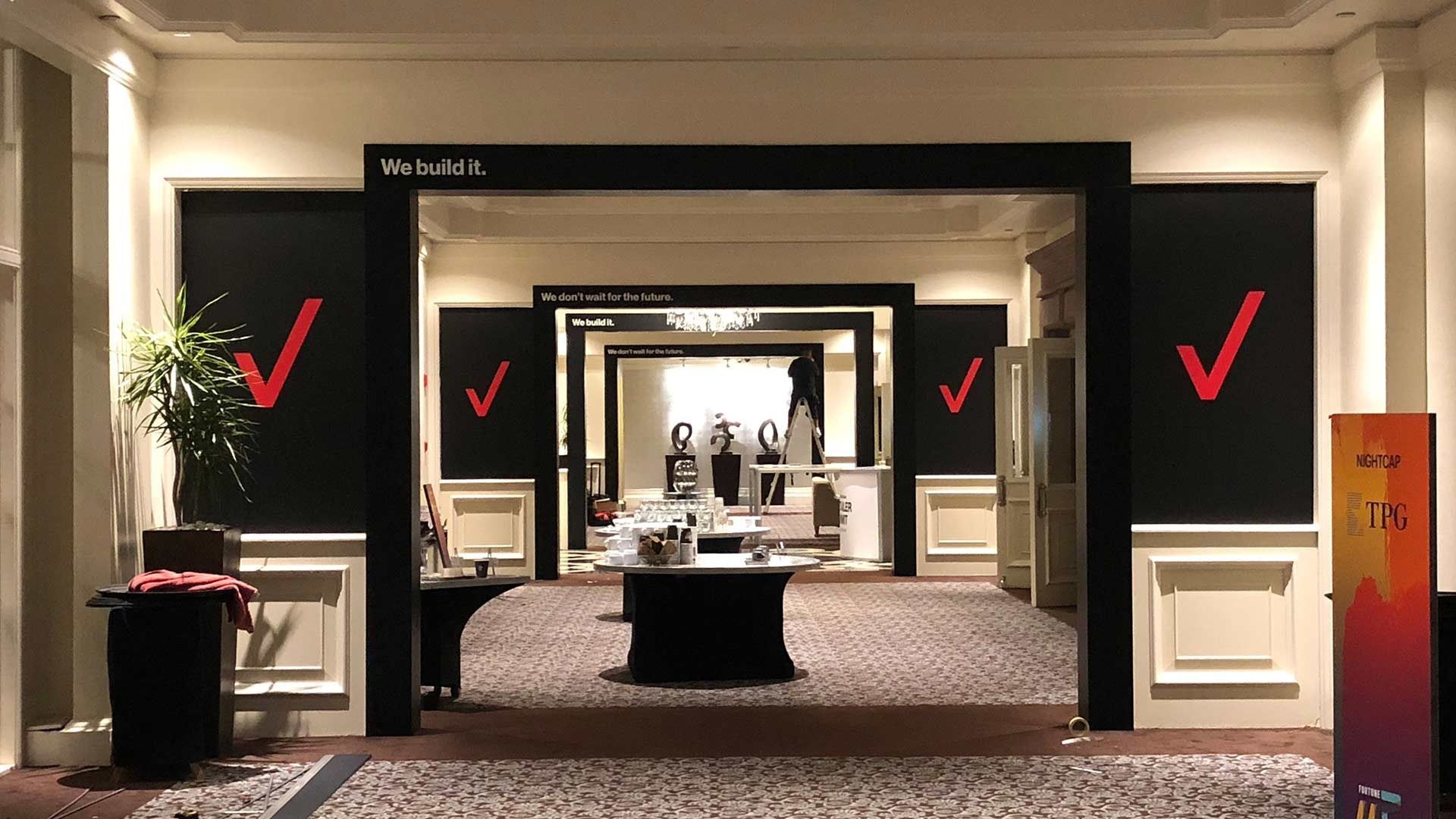 A hallway with repeating black rectangular arches and Verizon logo branding, featuring tables and a plant in the foreground.