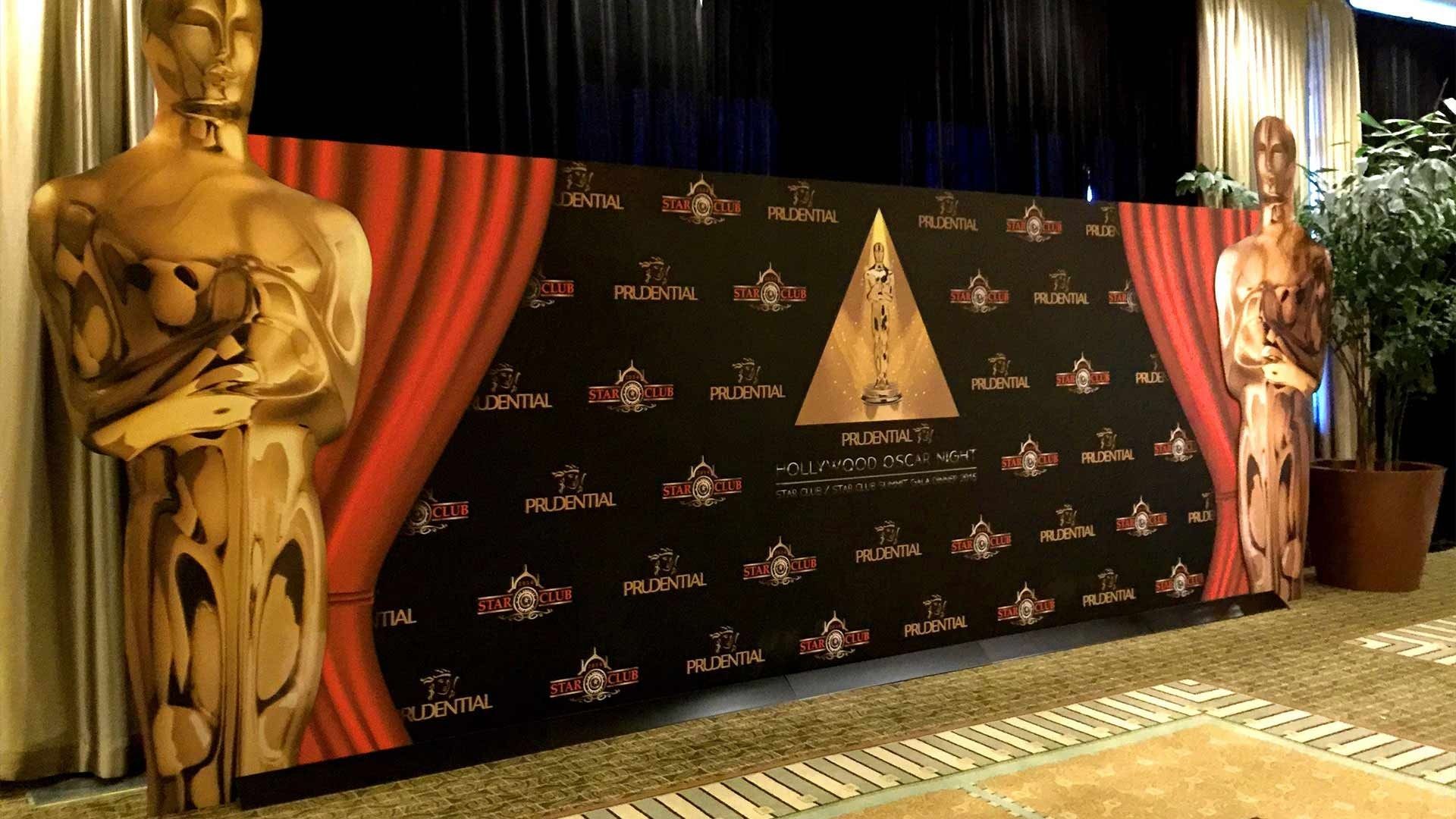 A step-and-repeat backdrop for an awards event, featuring red curtains, two gold Oscar-style statues, and a central emblem.