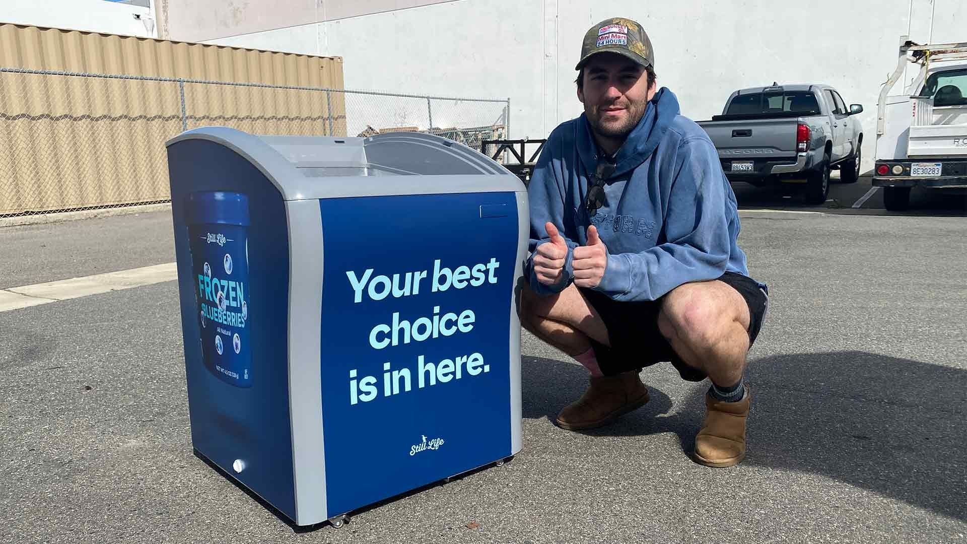 A person in a blue hoodie and cap squats next to a blue and gray marketing bin that says, 