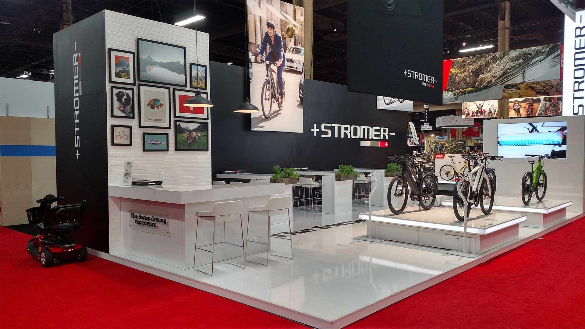 A modern trade show booth featuring Stromer branded signage, bicycle displays, a seating area, and framed wall artwork.
