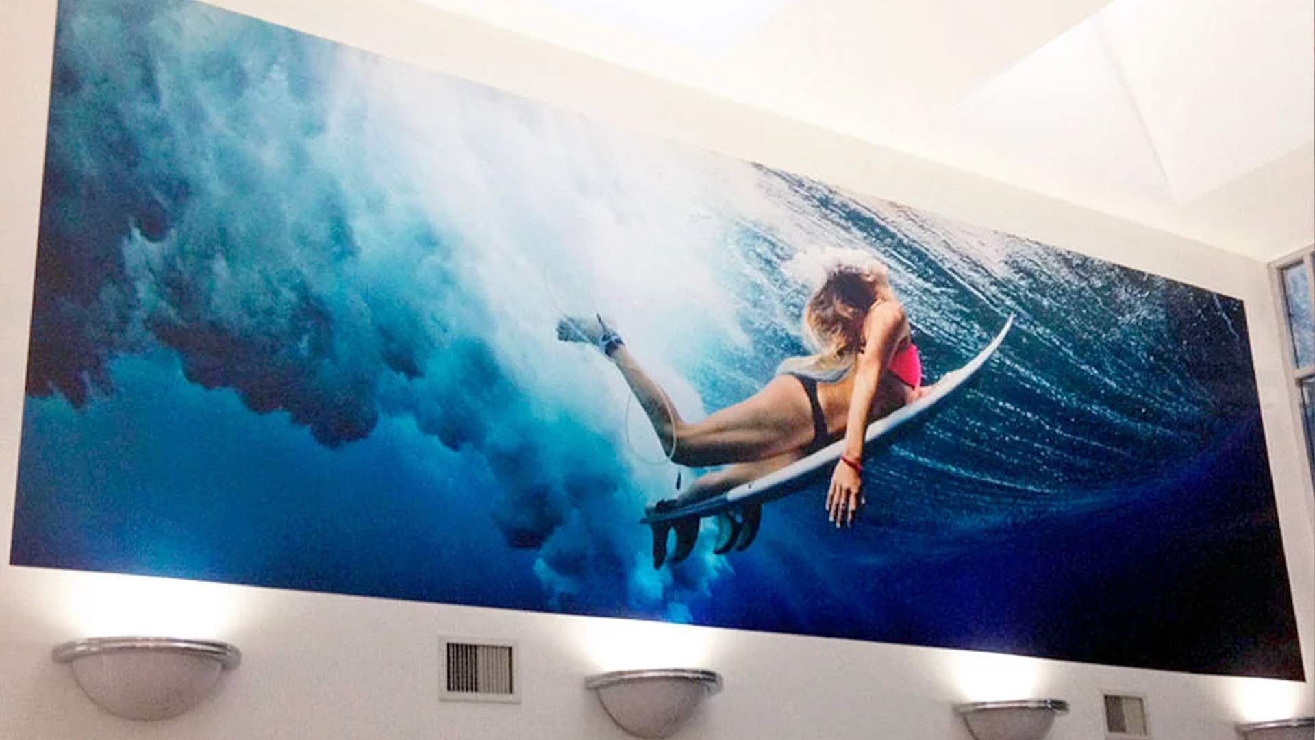 A large wall mural of a surfer riding inside a powerful, deep blue wave.