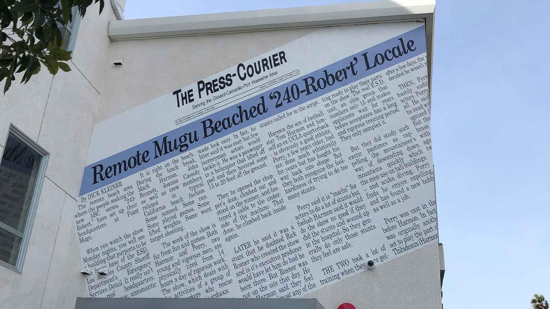 A mural on a white building wall displays a newspaper front page titled The Press-Courier, featuring the headline Mugu.