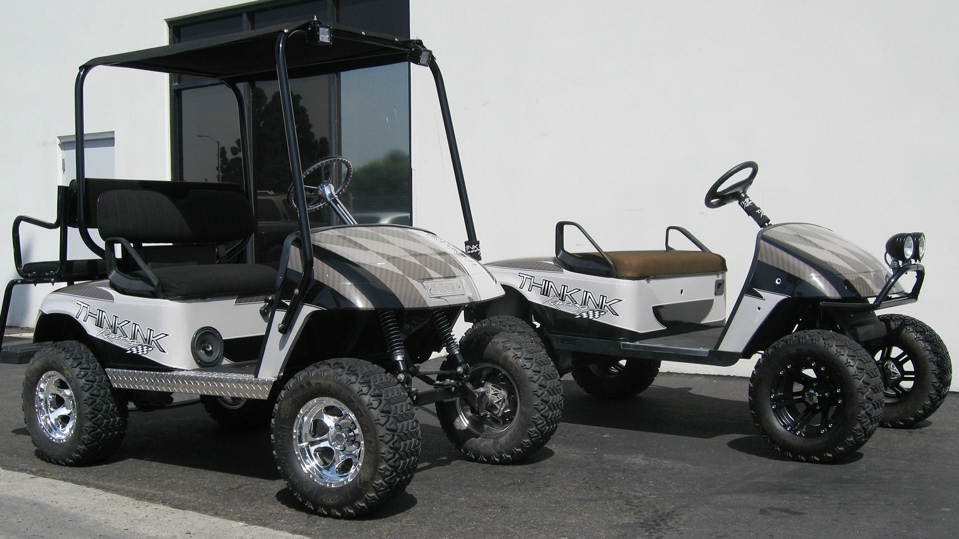 Two white golf carts with black accents and rugged tires parked side-by-side against a white building.