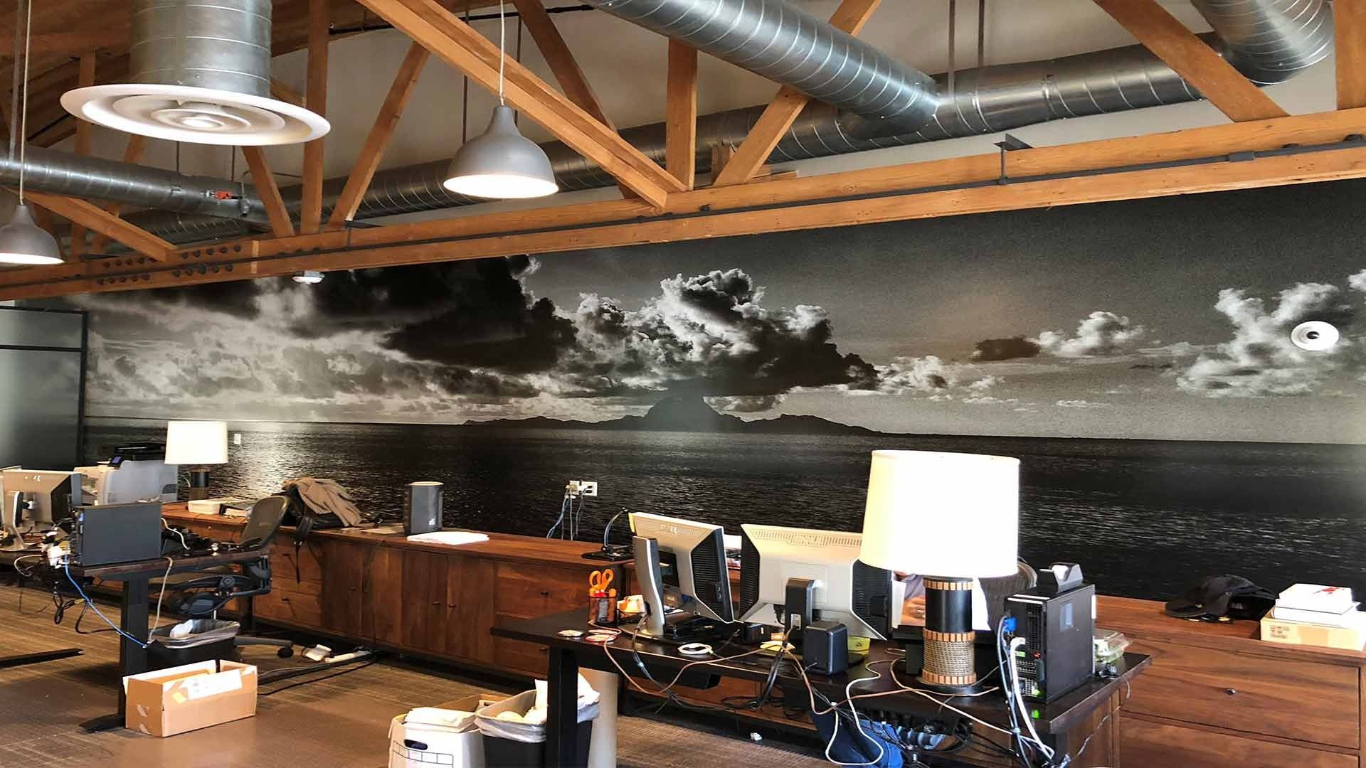 An office interior featuring a large black-and-white ocean mural on the wall, wooden beams, and desk workstations.