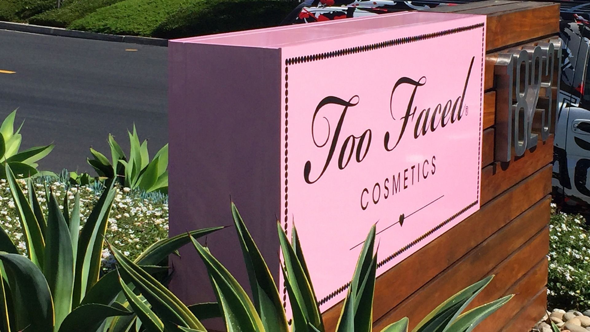 A pink Too Faced Cosmetics sign attached to a wooden pillar, surrounded by leafy plants.