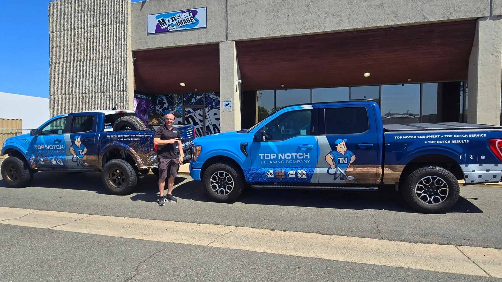 A person stands between two blue Top Notch Landscaping pickup trucks parked in front of a building on a sunny day.