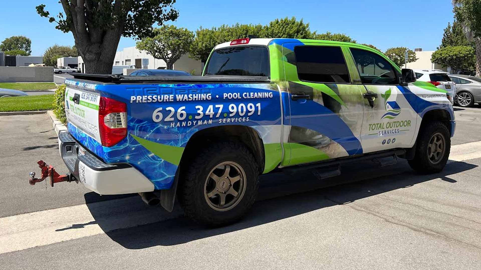 A blue, white, and green wrapped pickup truck parked in a lot, displaying a business phone number and logo on the side.