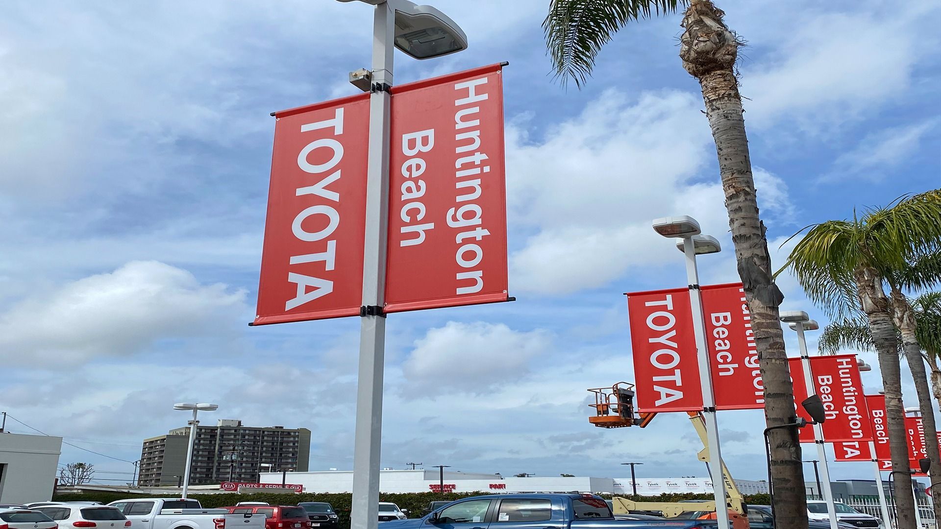 Red banners for Huntington Beach Toyota line a car dealership lot under a cloudy blue sky.