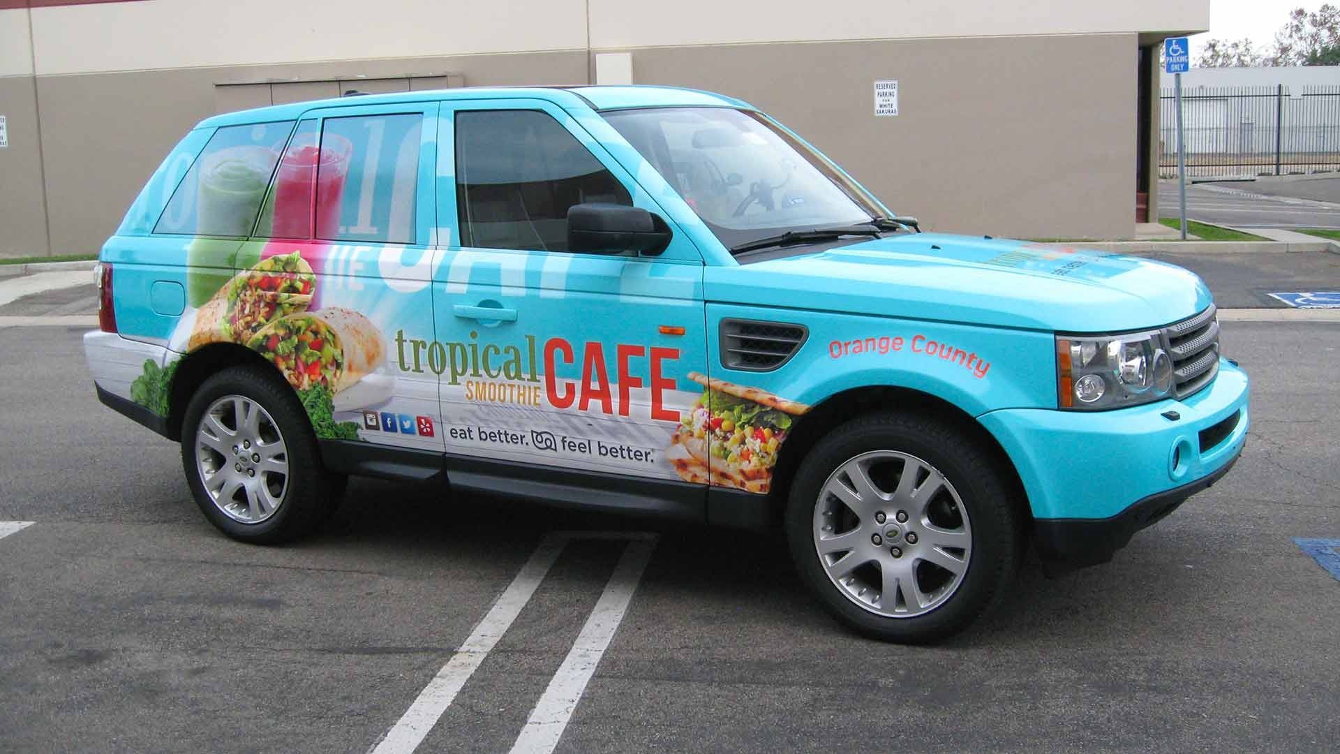 A light blue SUV parked in a lot, wrapped with Tropical Cafe branding featuring colorful food and drink graphics.