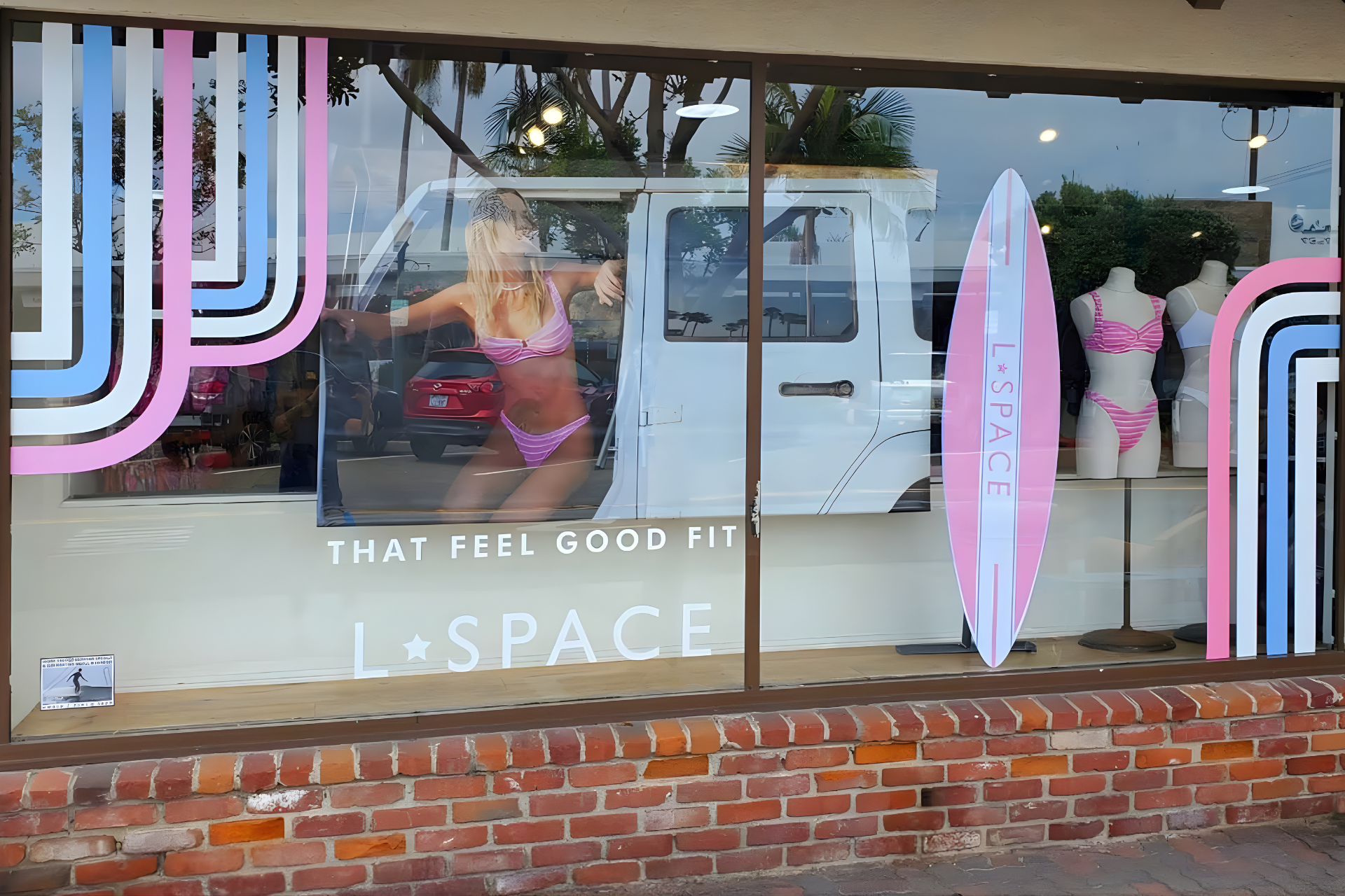 A storefront with white walls and fashion window displays behind a red car and a black sedan on a city street.