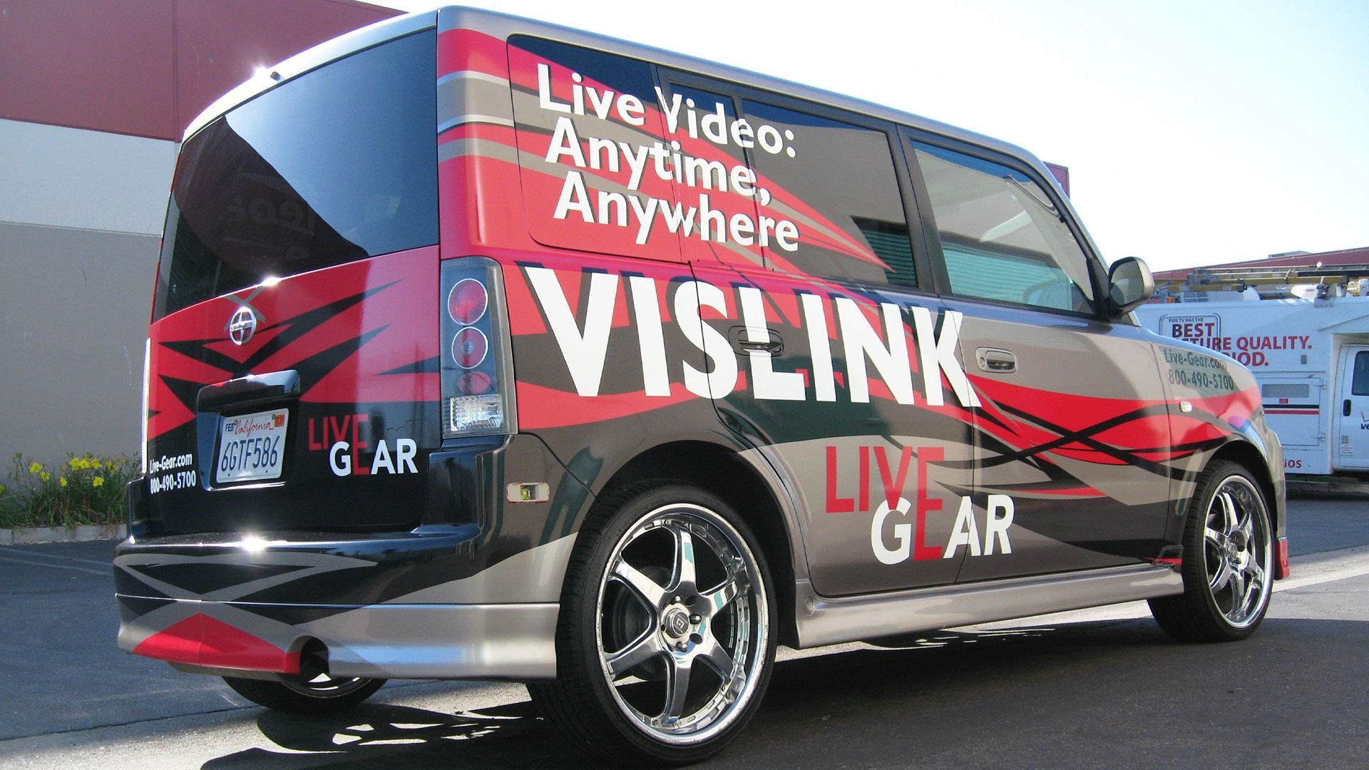 A Scion xB van wrapped in red, black, and grey branding for Vislink Live Gear parked outdoors.