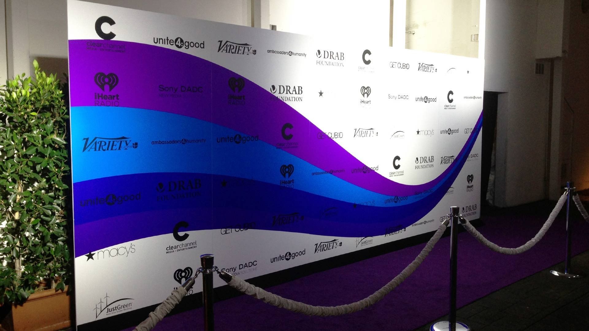 A step-and-repeat media backdrop with purple and blue waves, sponsor logos, and a purple carpet with velvet rope stanchions.