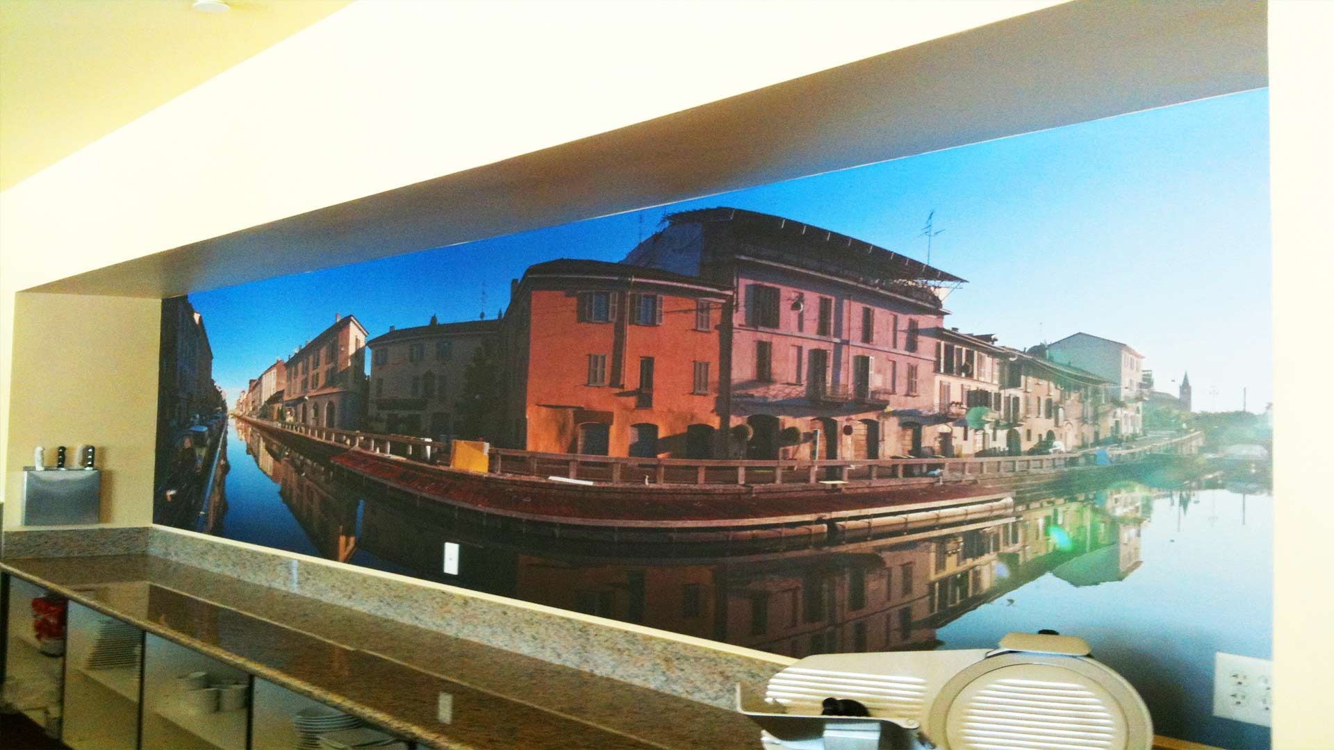 A mural of a canal-side town with multi-colored buildings reflects into calm water on a long wall above a countertop.