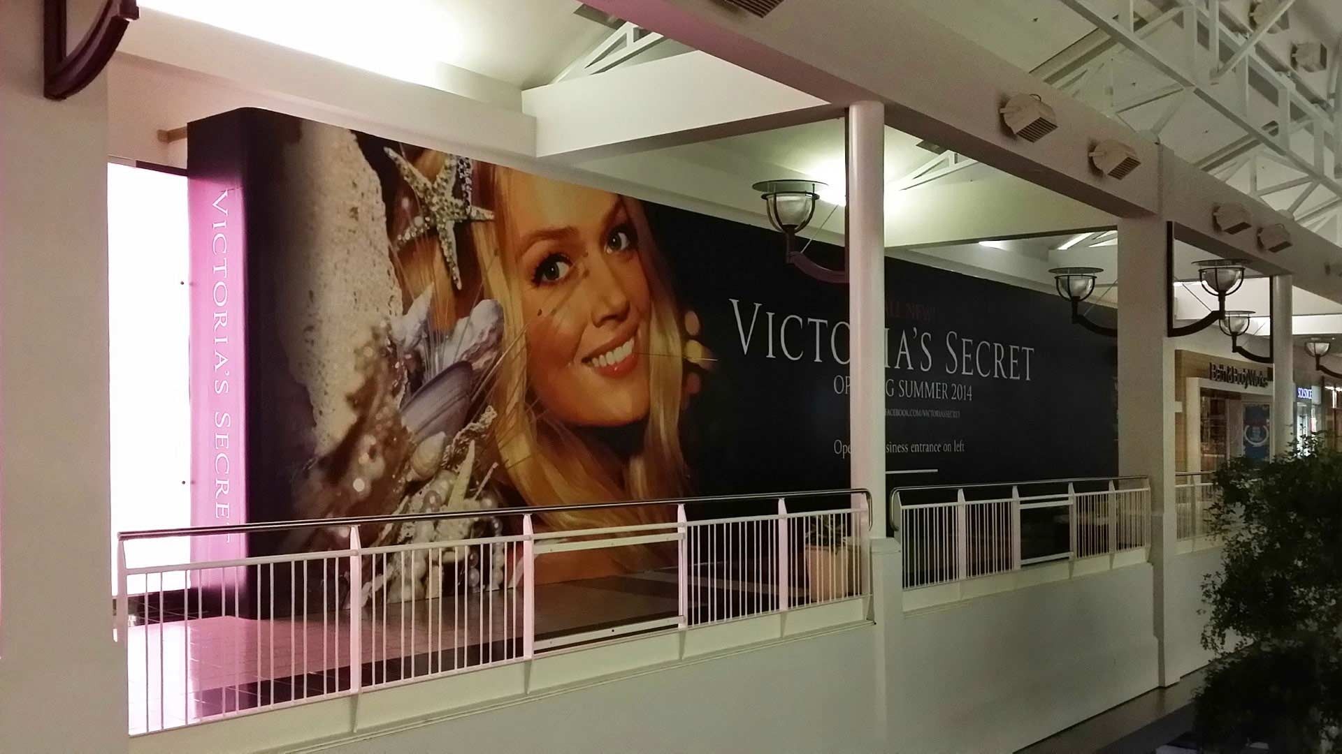 A Victoria's Secret advertisement banner featuring a woman on a wall in a shopping mall.