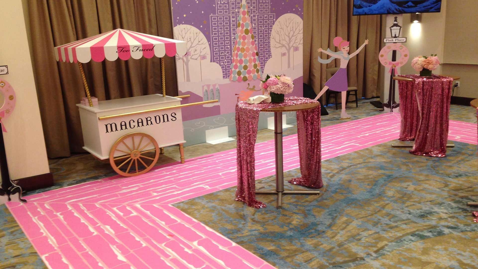 A pink-themed party setup with a macaron cart, tall cocktail tables draped in sequined cloths, and a decorative backdrop.